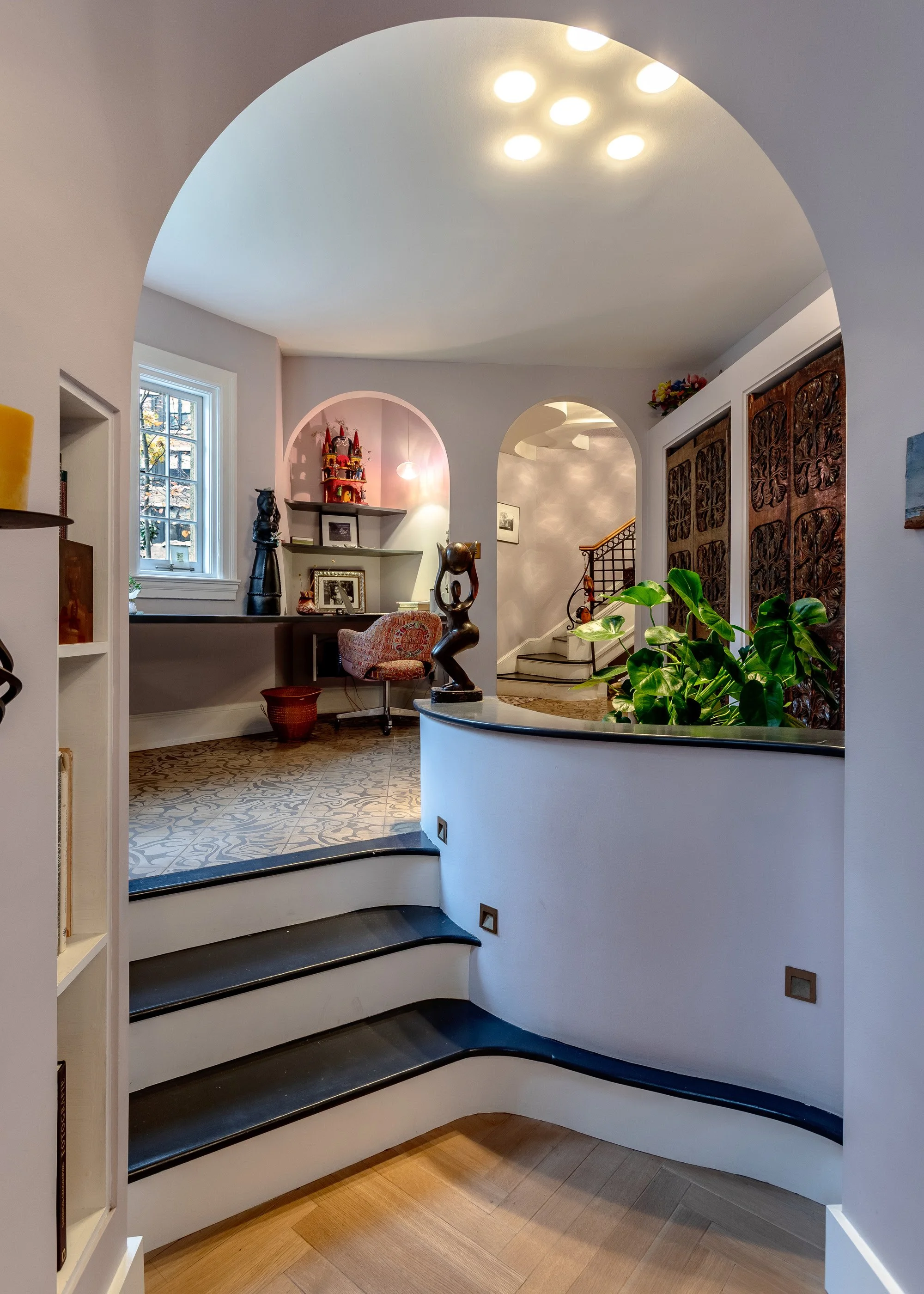 Interior view of a home with a staircase, a curved ledge, decorative sculptures, a window, and patterned tiles in the background.