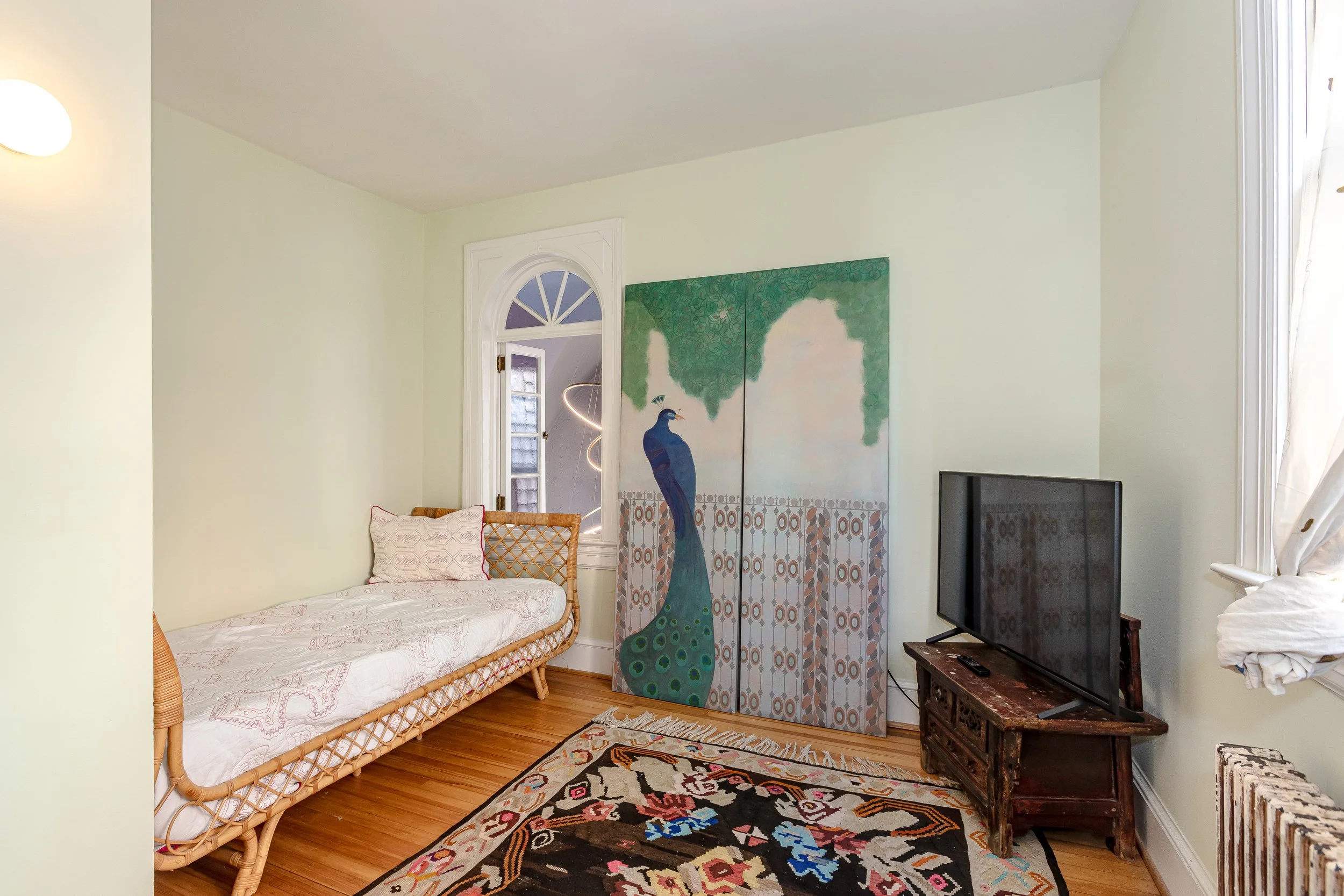A cozy room with a rattan daybed with patterned white and pink bedding and a pillow, honey-colored wooden flooring, a colorful peacock artwork on a foldable screen, a patterned rug, an old wooden TV stand with a flat-screen TV, a small remote, a wind