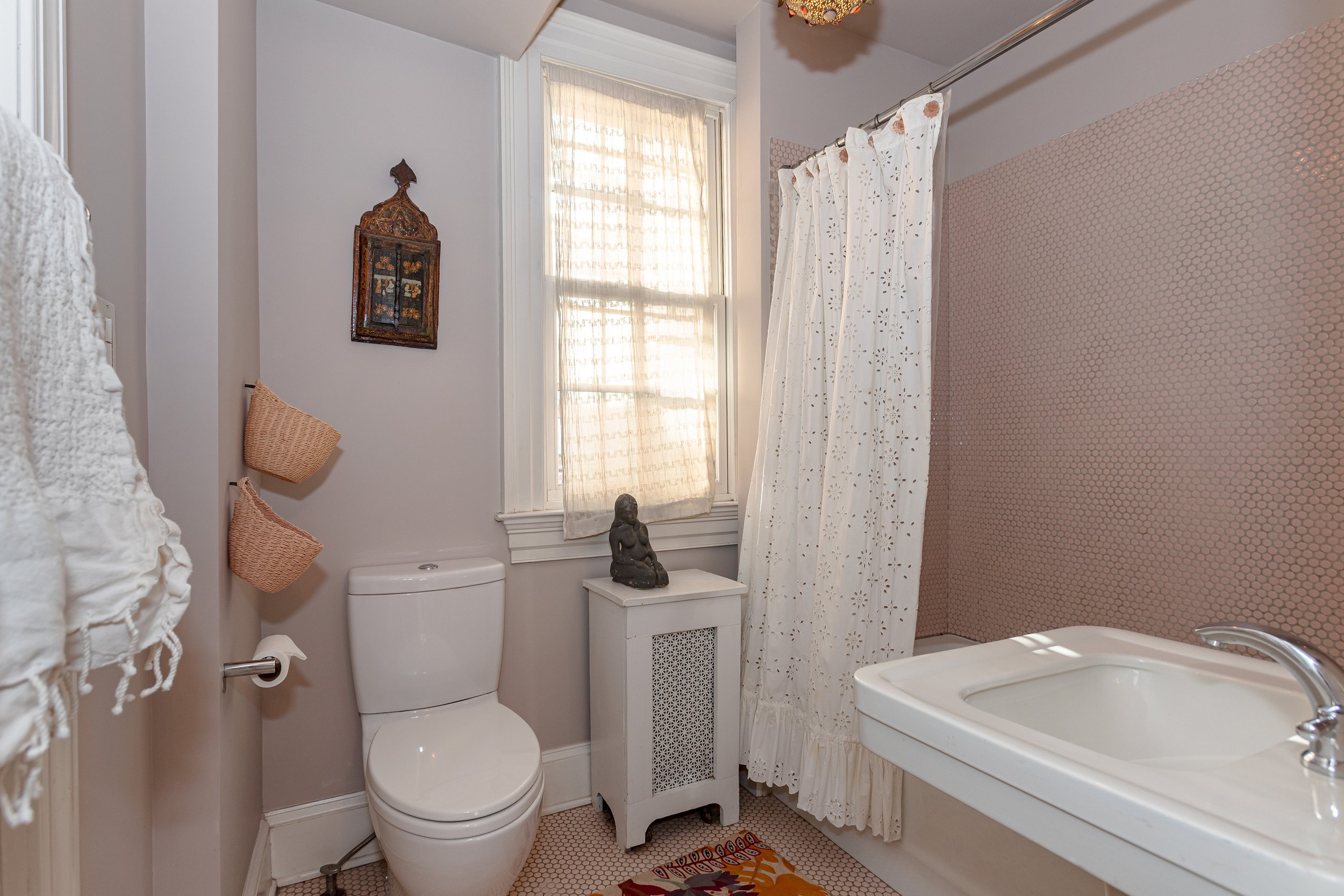 A small bathroom with a toilet, a white sink, and a window with white curtains. There is a decorative wall hanging above the toilet and two hanging woven baskets on the side. A small statue is on a white cabinet near the window. The wall behind the b