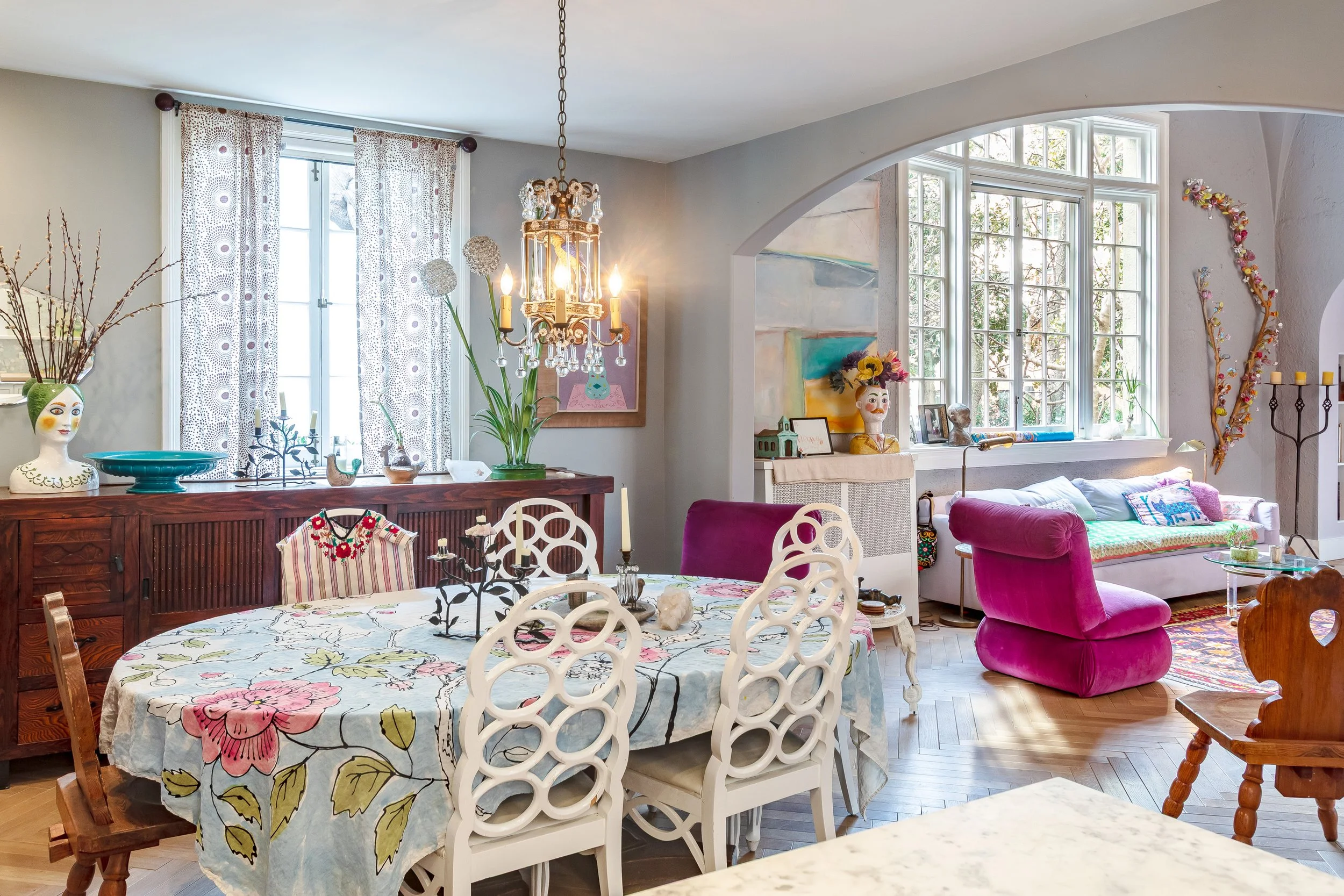 A living and dining room with a round floral dining table, eclectic chairs, large windows, colorful artwork, a chandelier, and cozy seating including hot pink velvet chairs and a sofa with colorful pillows, decorated with art and decorative objects.
