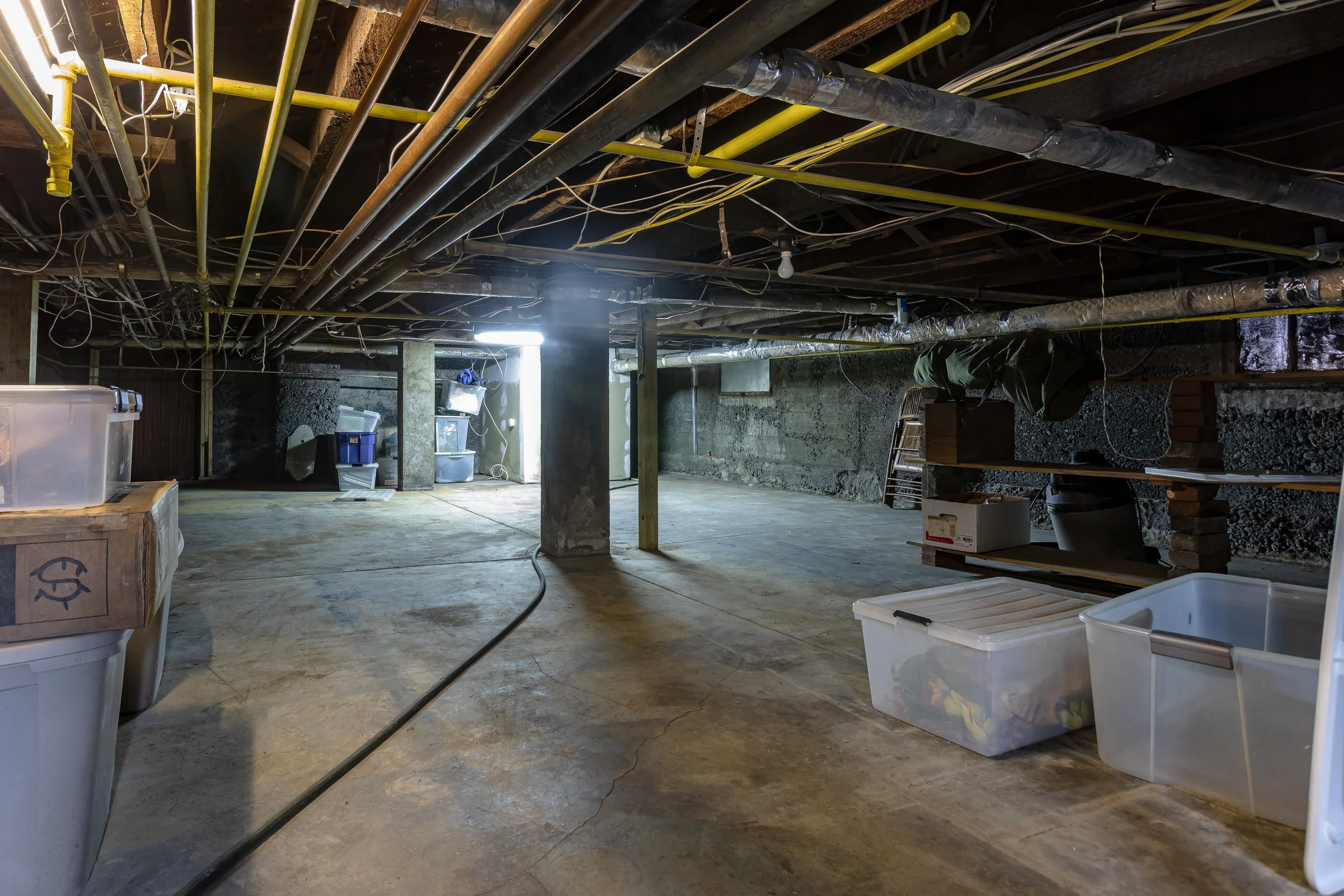 Empty basement with exposed ceiling pipes, concrete walls, and scattered plastic storage bins.