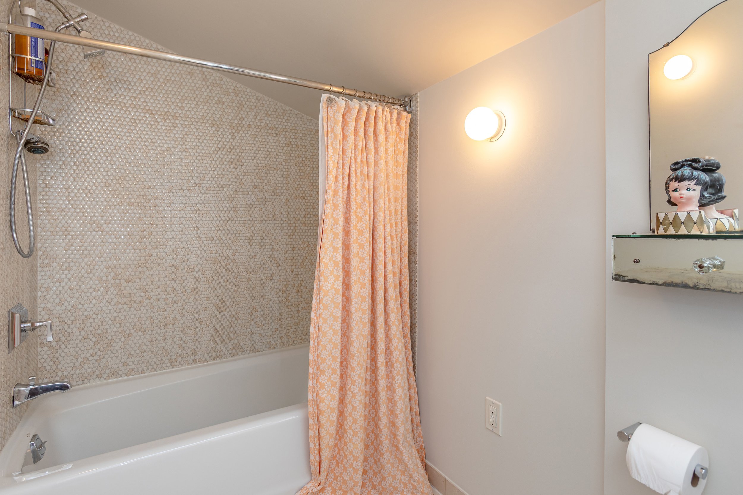 Bathroom with a bathtub, shower, shower curtain, wall-mounted lights, mirror, decorative doll, and toilet paper holder.