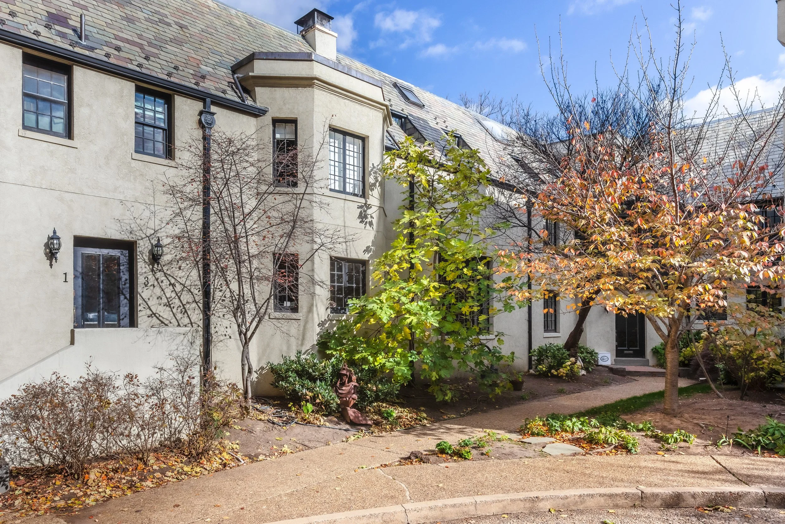 A residential building with beige stucco walls, multiple windows, and a multi-gabled slate roof. The front yard has seasonal trees and shrubs, a pathway, and a small statue among the plants. The sky is partly cloudy with sunlight illuminating the sce