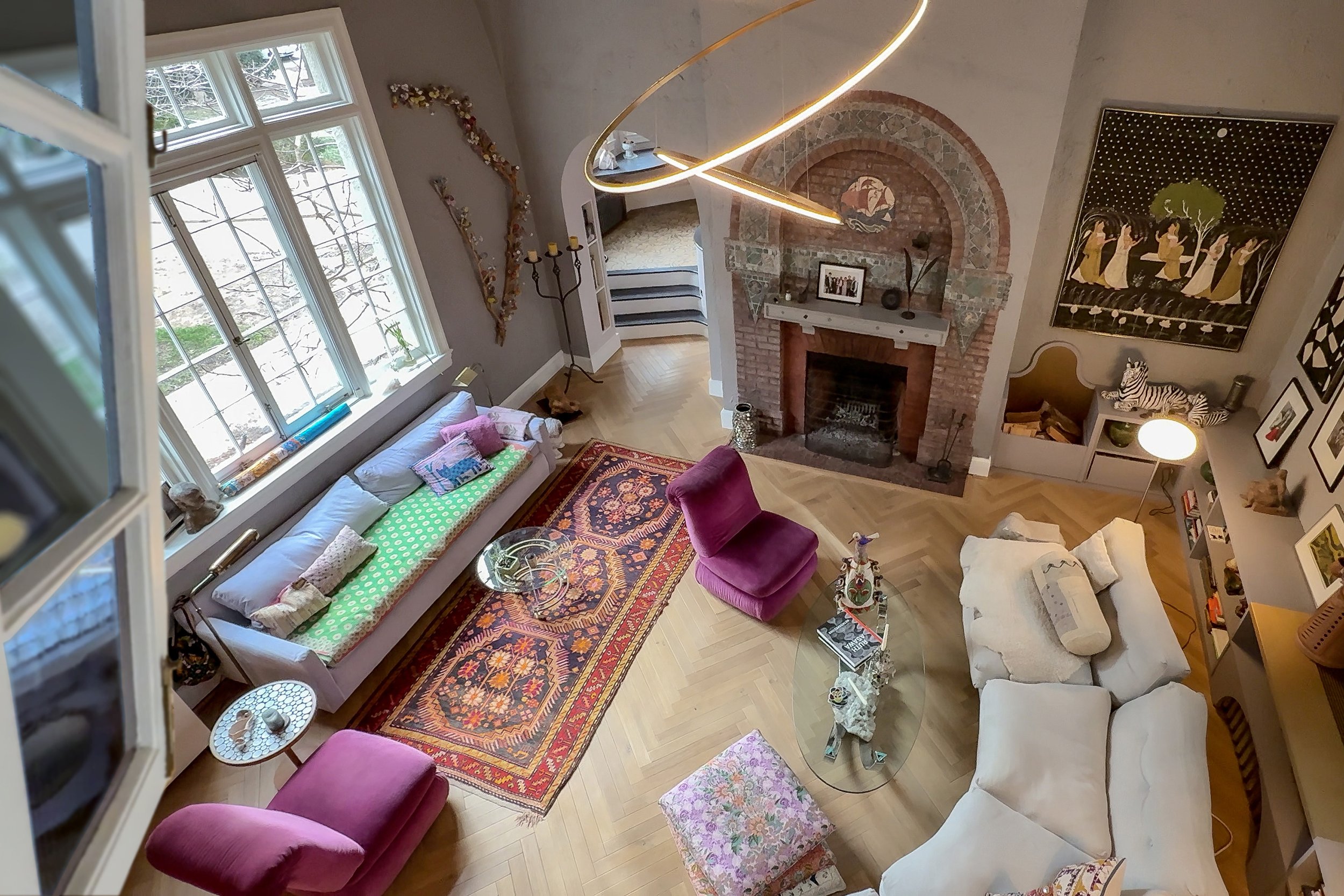 A cozy living room with a large window, a white sofa with colorful cushions, two pink armchairs, a glass coffee table, a patterned area rug, a brick fireplace, modern chandelier, wall art, and various decorative items.