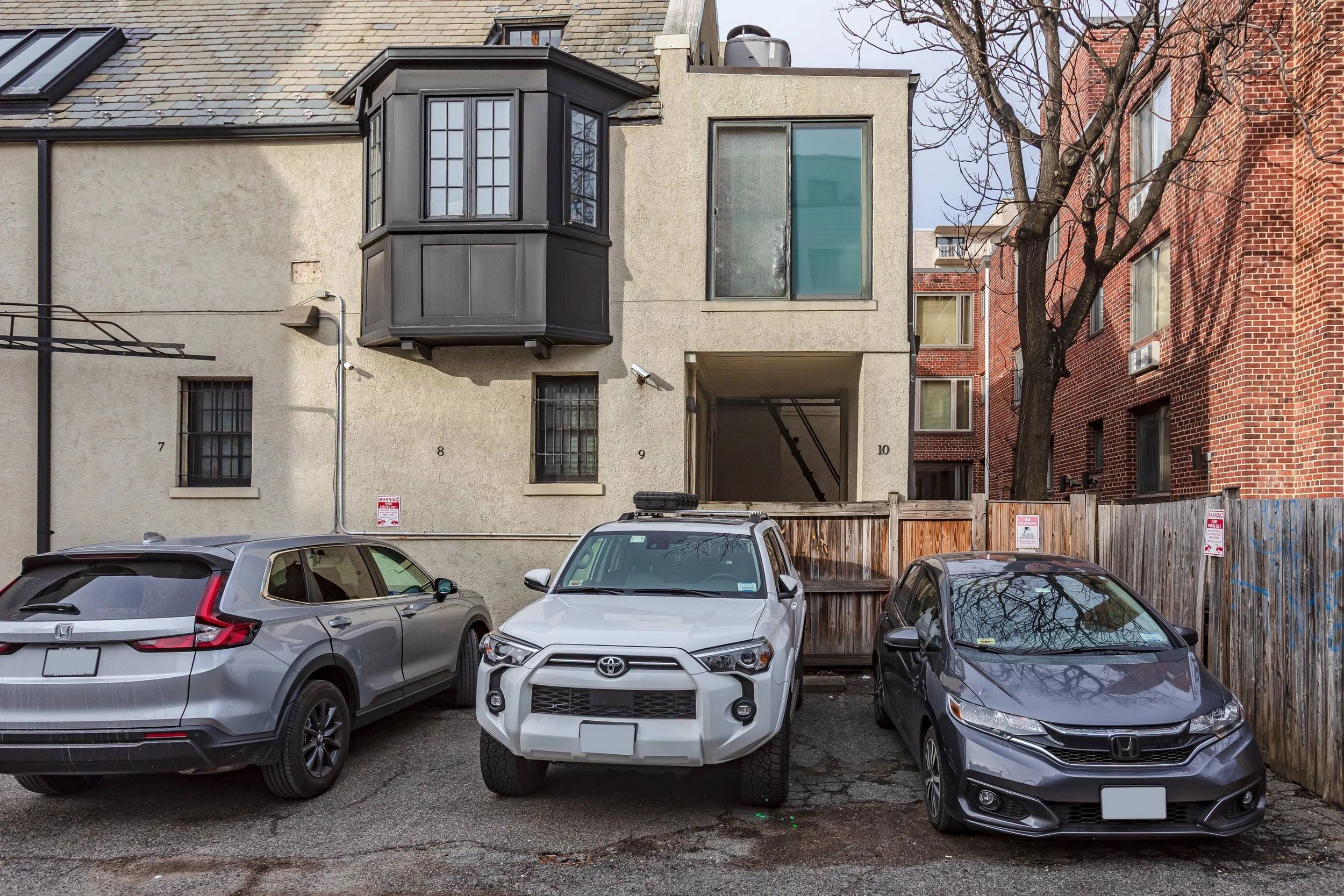 Three parked cars in front of a multi-story building with a beige facade, black bay window, and a small tree on the right side.