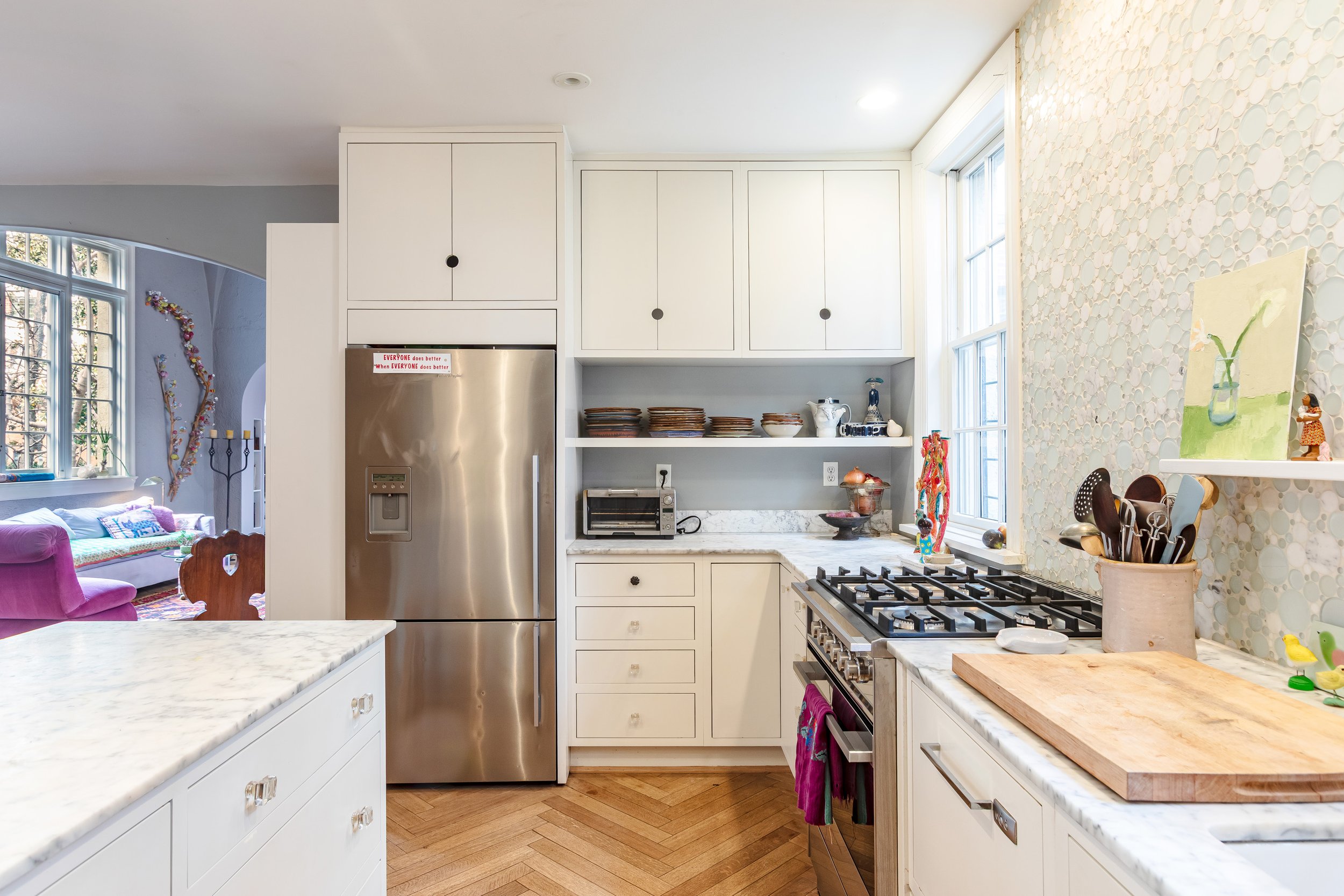 Bright kitchen with white cabinets, marble countertops, a stainless steel refrigerator, a gas stove, and open shelving with plates and decorative items. A large window provides natural light.