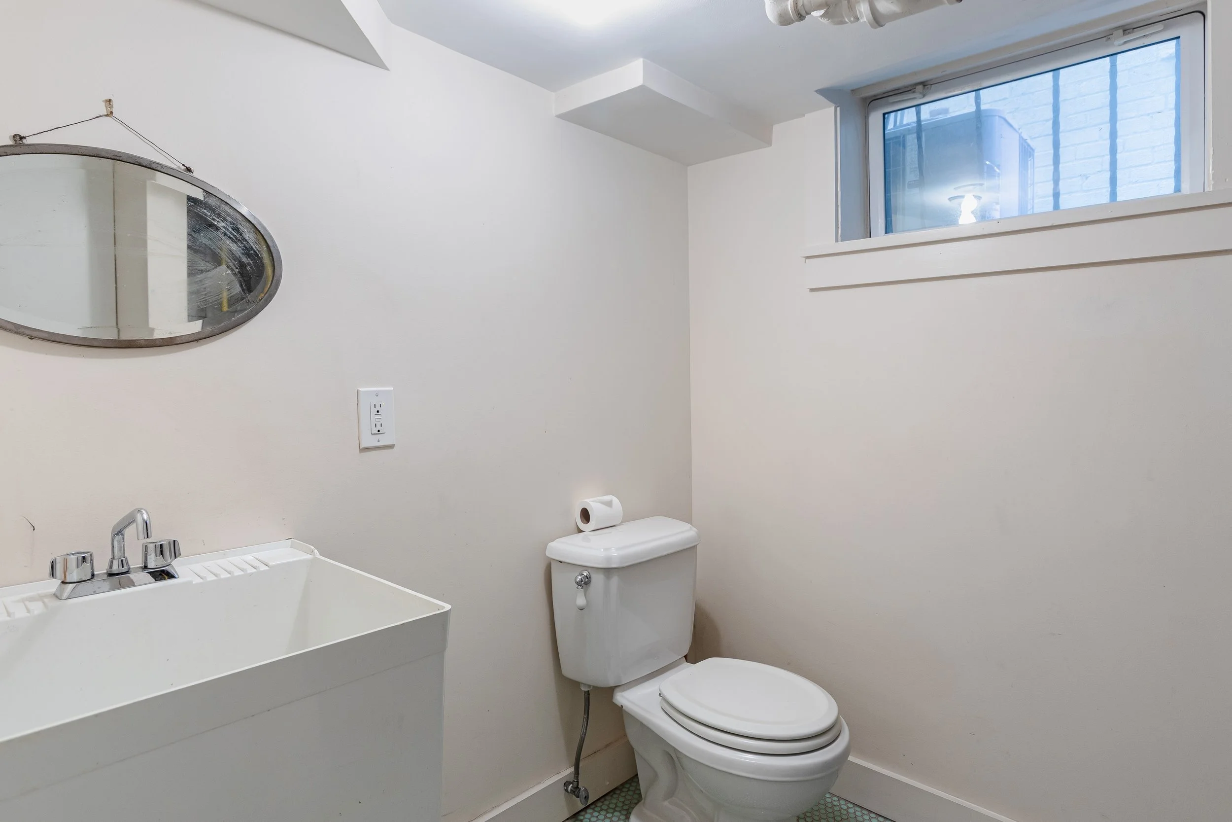 Small bathroom with white walls, a toilet with a roll of toilet paper on top, a small window, a white utility sink, and a mirror on the wall.