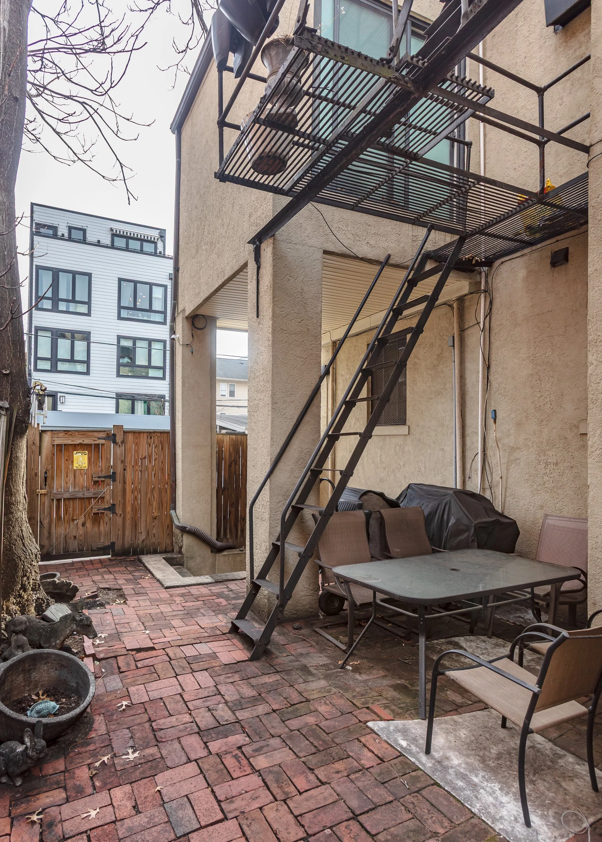A small outdoor patio with brick flooring and a beige stucco building wall in the background. There is a metal staircase leading to a second-floor balcony. The patio has outdoor furniture, including a glass-top table with four chairs, a covered grill