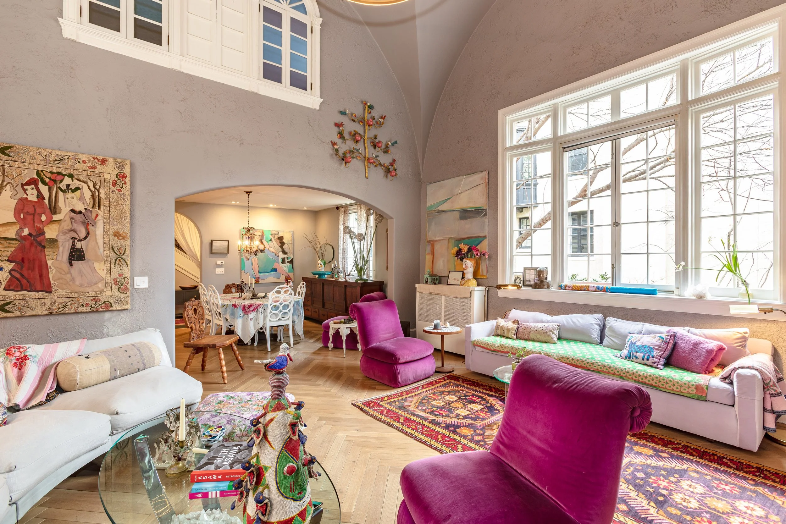 A colorful living room with large windows, pink and purple chairs, a white sofa with pillows, paintings on the walls, and a view into a dining area with a chandelier and a table with chairs.
