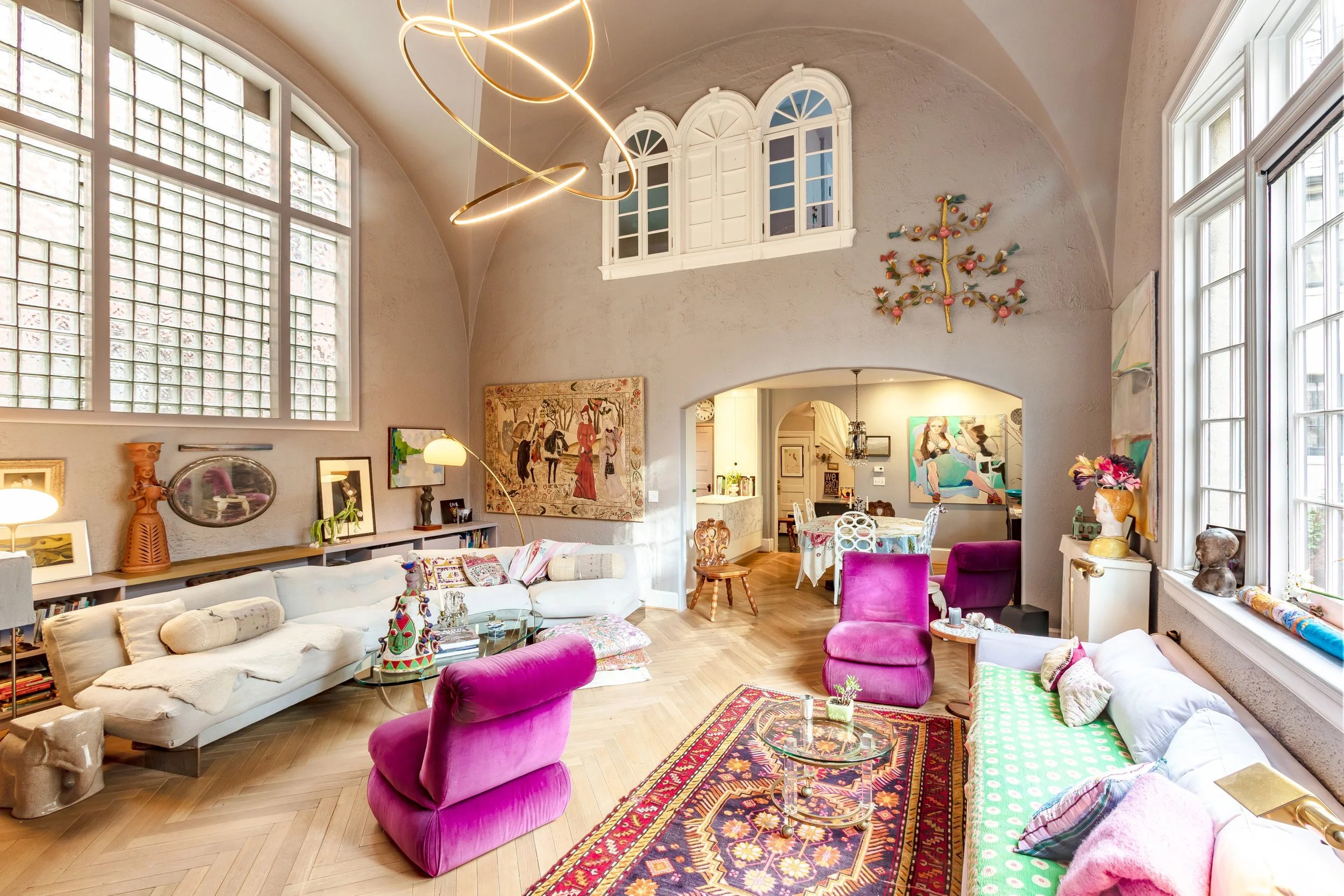 Spacious living room with large windows, eclectic furniture including white and bright pink chairs, and art on the walls, with a view into the dining area.