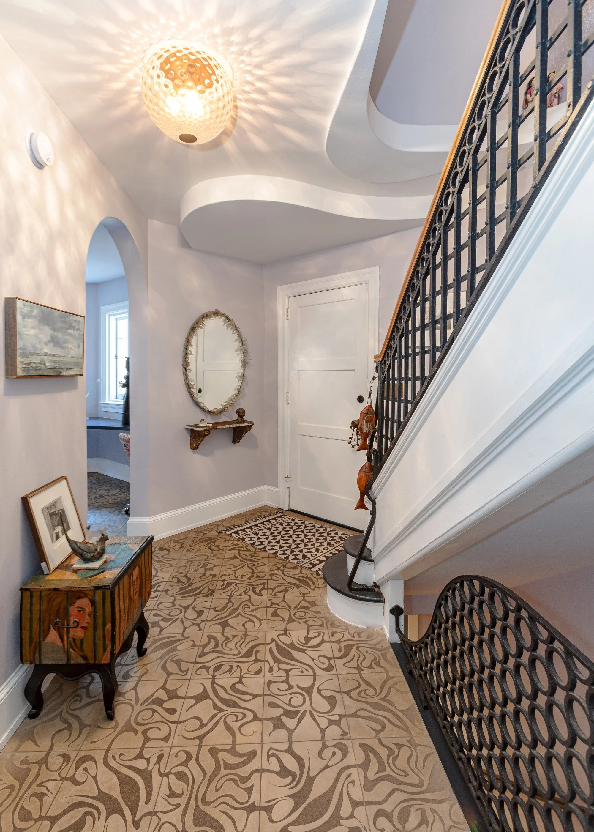 Interior of a home entryway with patterned tile flooring, a white door, a staircase with black iron railing, a small wooden side table with a painting of a woman, a mirror on the wall, and decorative elements like a woven light fixture and artwork.