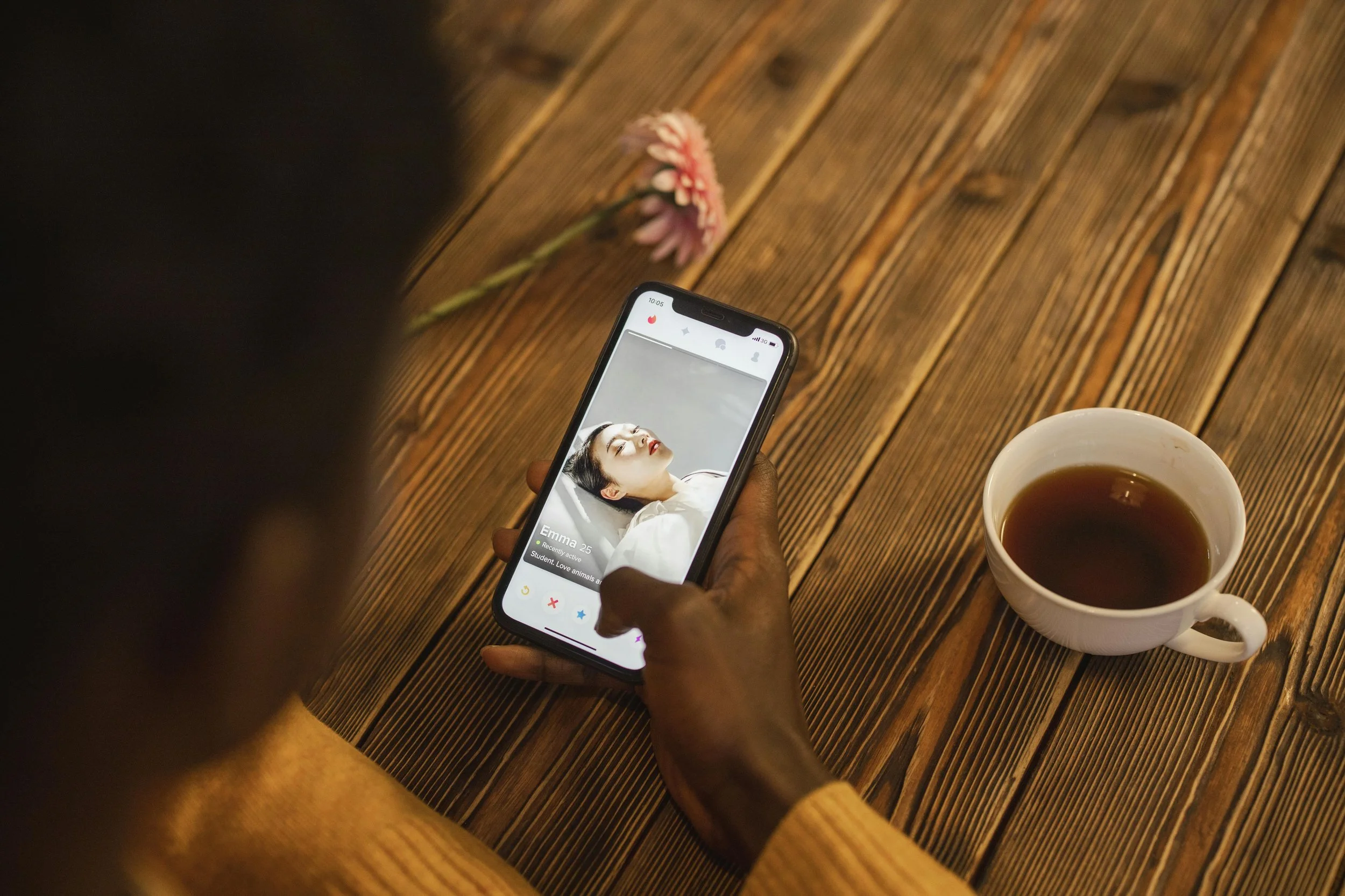 Person holding a smartphone showing a dating app profile of a woman named Emma, 25, with a cup of tea on a wooden table, and a pink flower in the background.