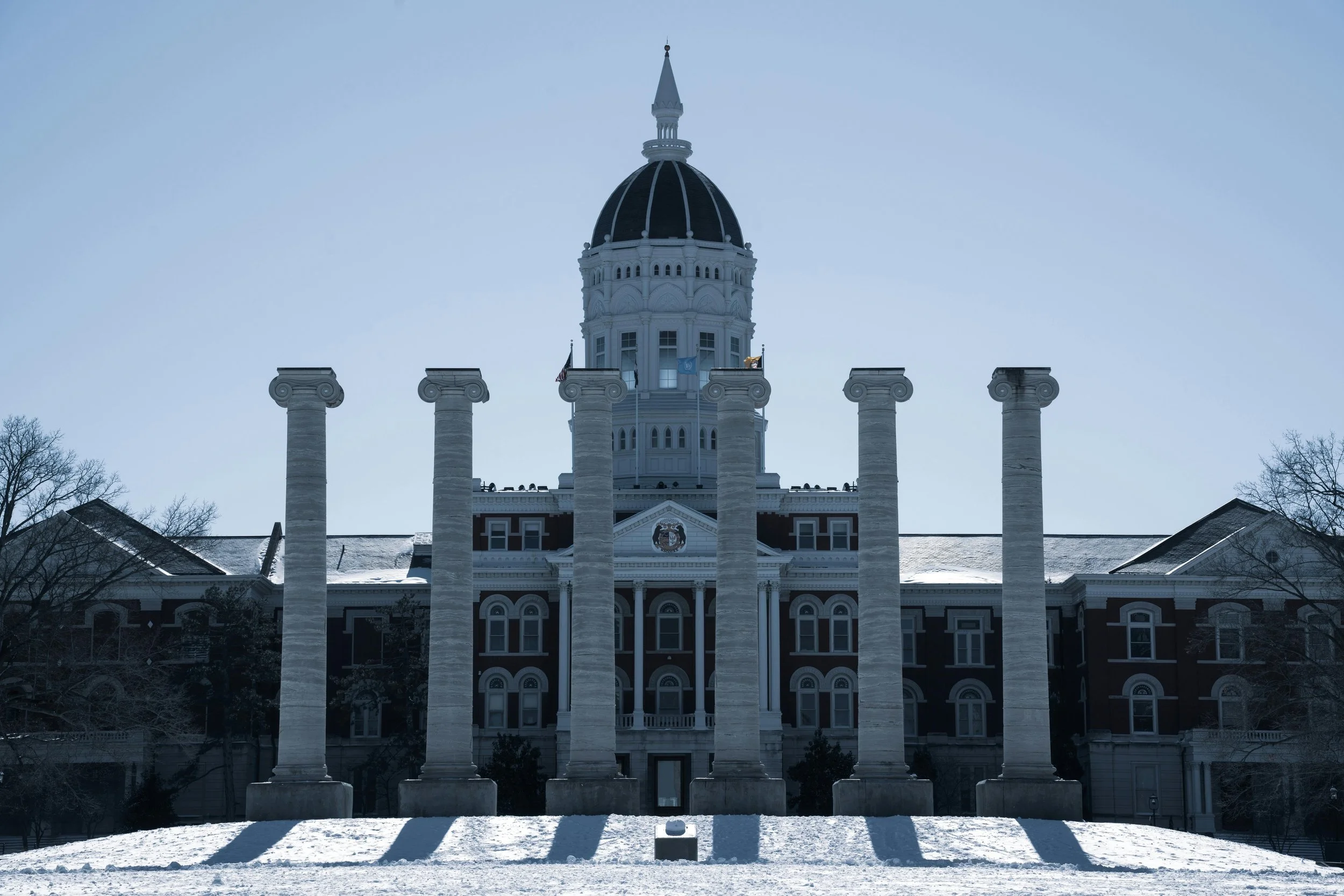 Historic government building with six tall columns in front, snow on ground, trees to the sides, and a dome on top of the building against a clear sky.