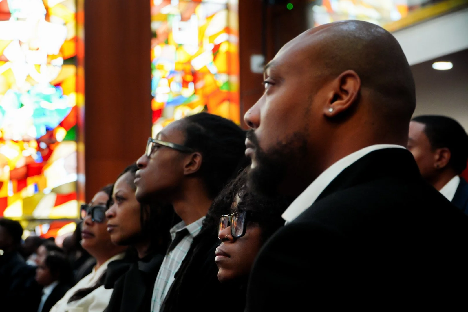 People attending a religious service or ceremony inside a church with colorful stained glass windows in the background.
