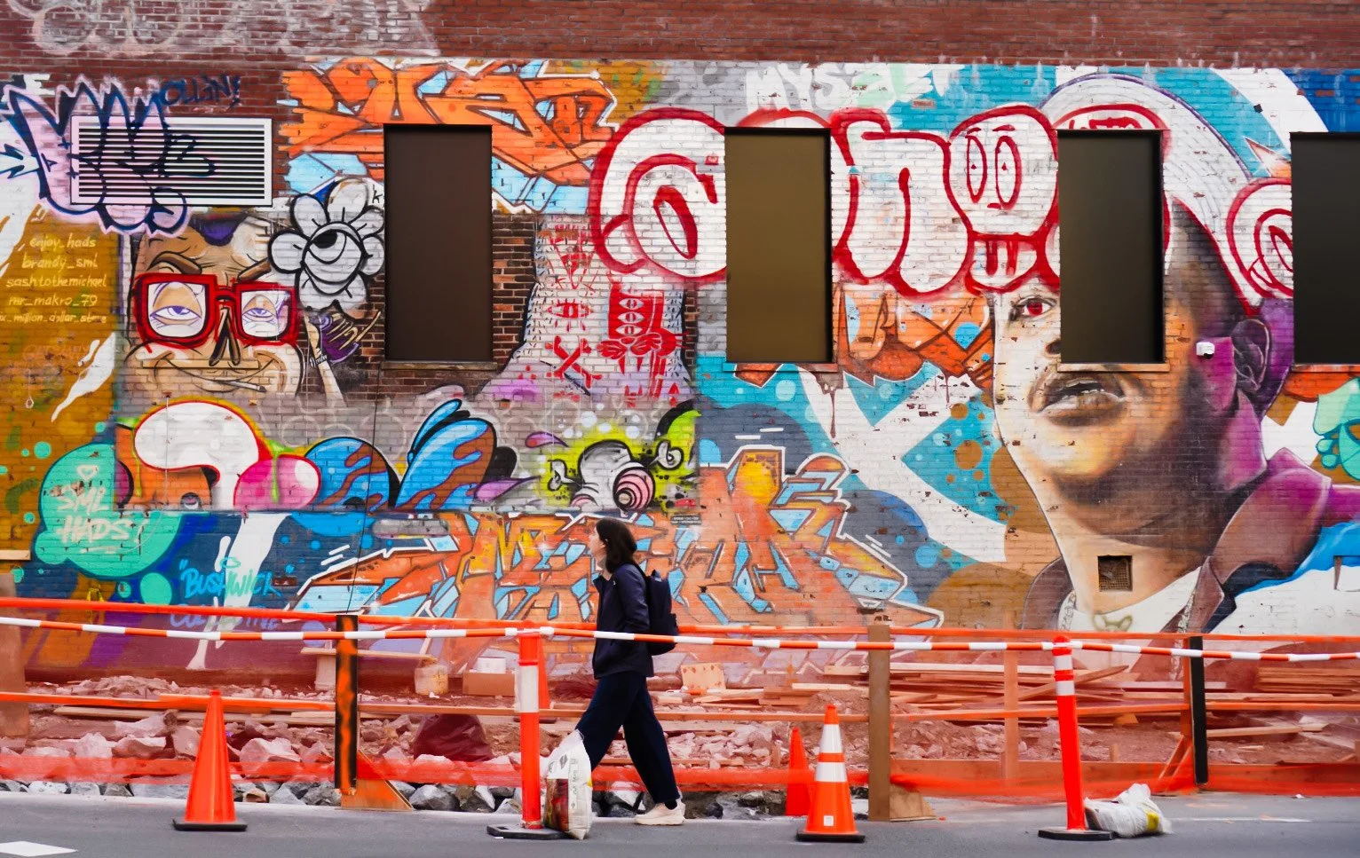 A vibrant street mural with graffiti art, including abstract characters and a woman's face, on a brick wall. A person with a backpack walks past orange construction cones and barriers in the foreground.
