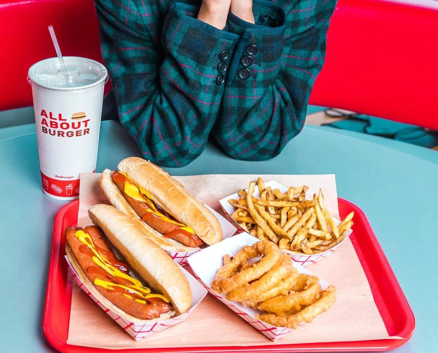 Fast food meal on a red tray, including two hot dogs with mustard, a side of French fries, fried onion rings, and a large soft drink in a white cup labeled "All About Burger" on a table with a teal surface, with a person in a green plaid jacket sitting behind.