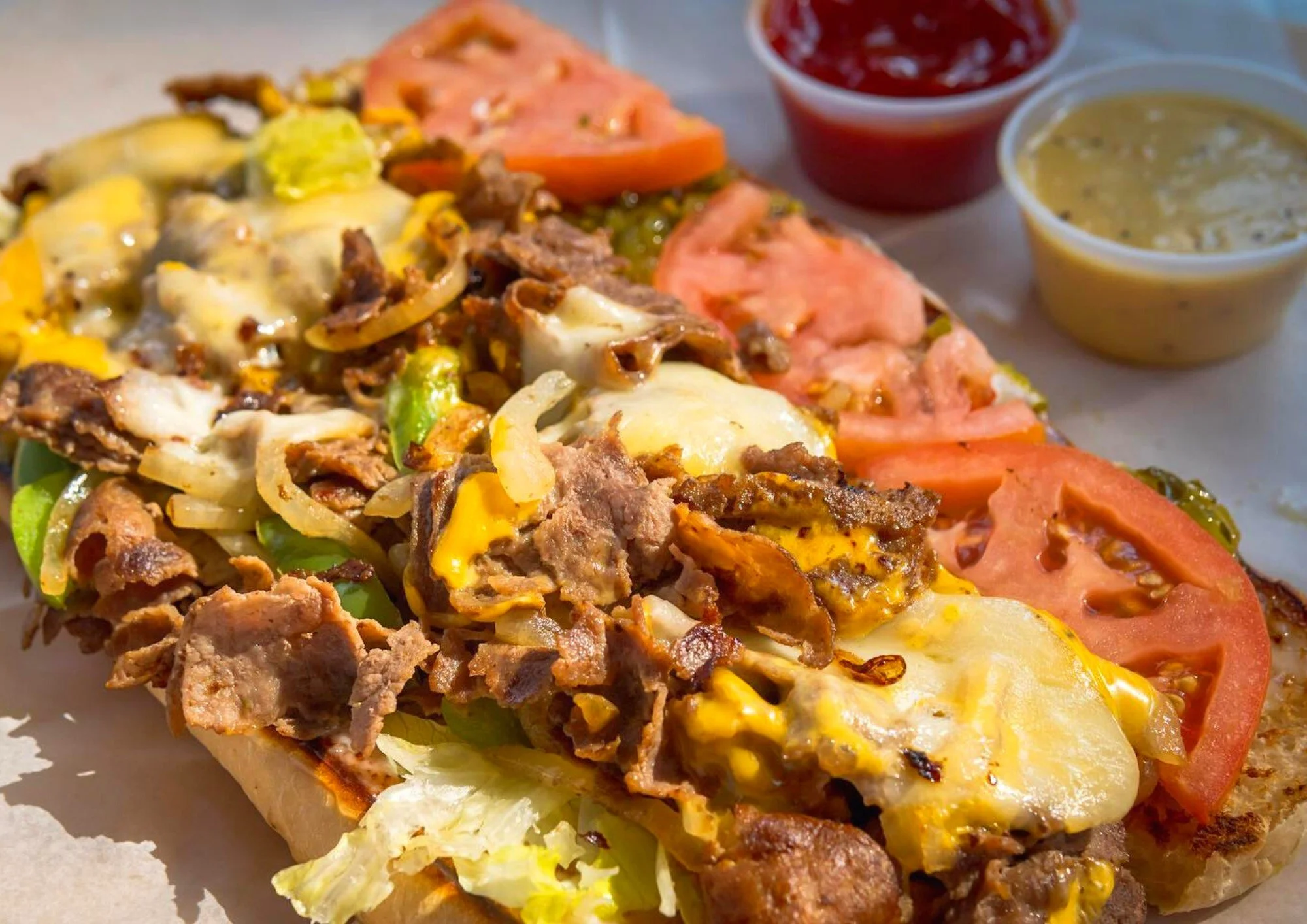 Close-up of a loaded Philly cheesesteak sandwich with melted cheese, sliced tomatoes, and condiments on the side.
