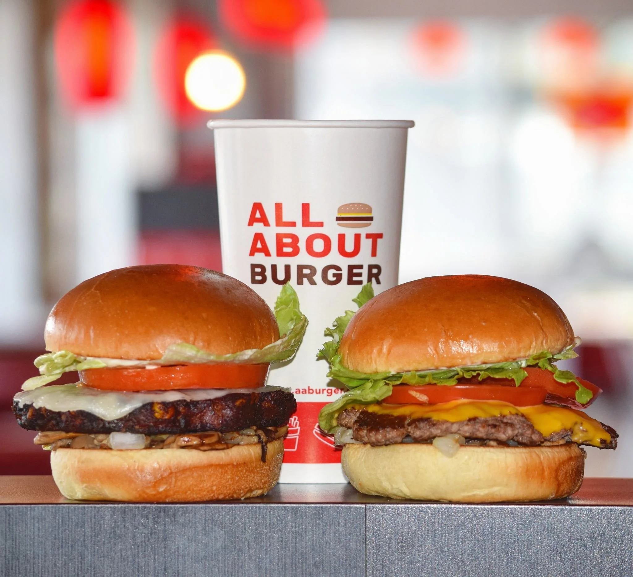 Two cheeseburgers with lettuce, tomato, cheese, and beef patties, and a paper cup with the label 'All About Burger' in the background.