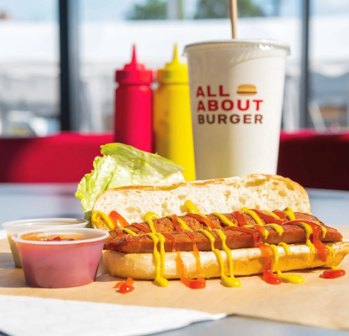 Hot dog with mustard and ketchup, lettuce, side sauces, a drink from All About Burger, and condiment bottles on a table.