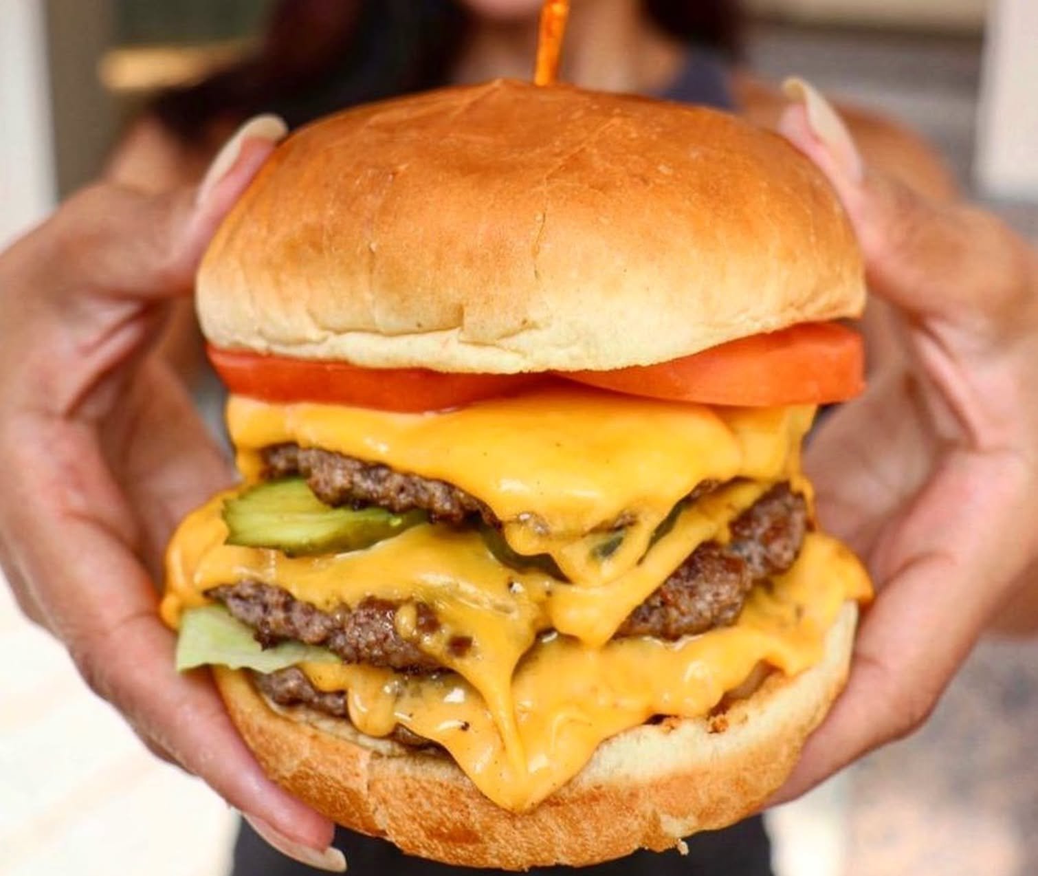 Close-up of a large cheeseburger with cheddar cheese, tomato, pickles, and beef patties, held with hands.