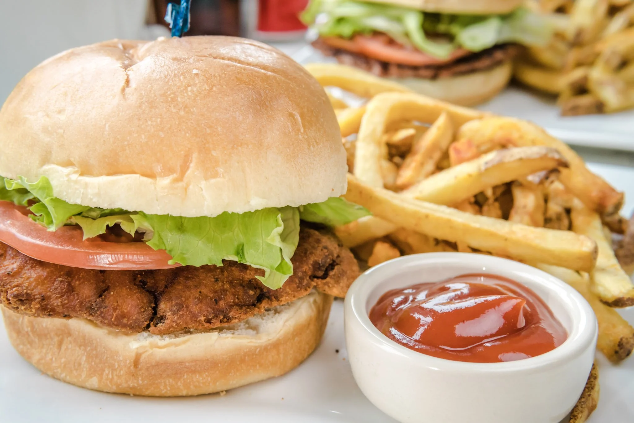 A crispy fried chicken sandwich with lettuce, tomato, and pickles in a bun, served with French fries and a small dish of ketchup.