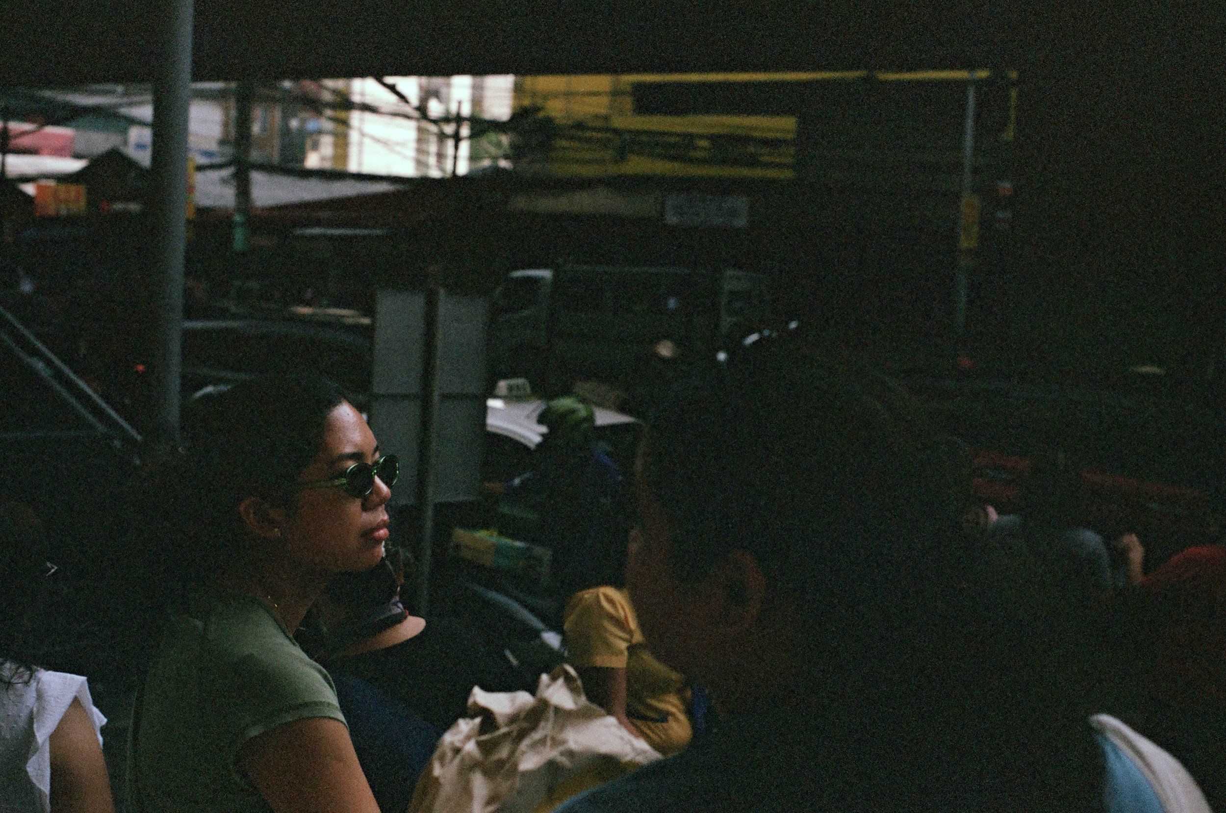 A woman wearing sunglasses sitting on a bus or train with other passengers, dark interior, city street visible outside
