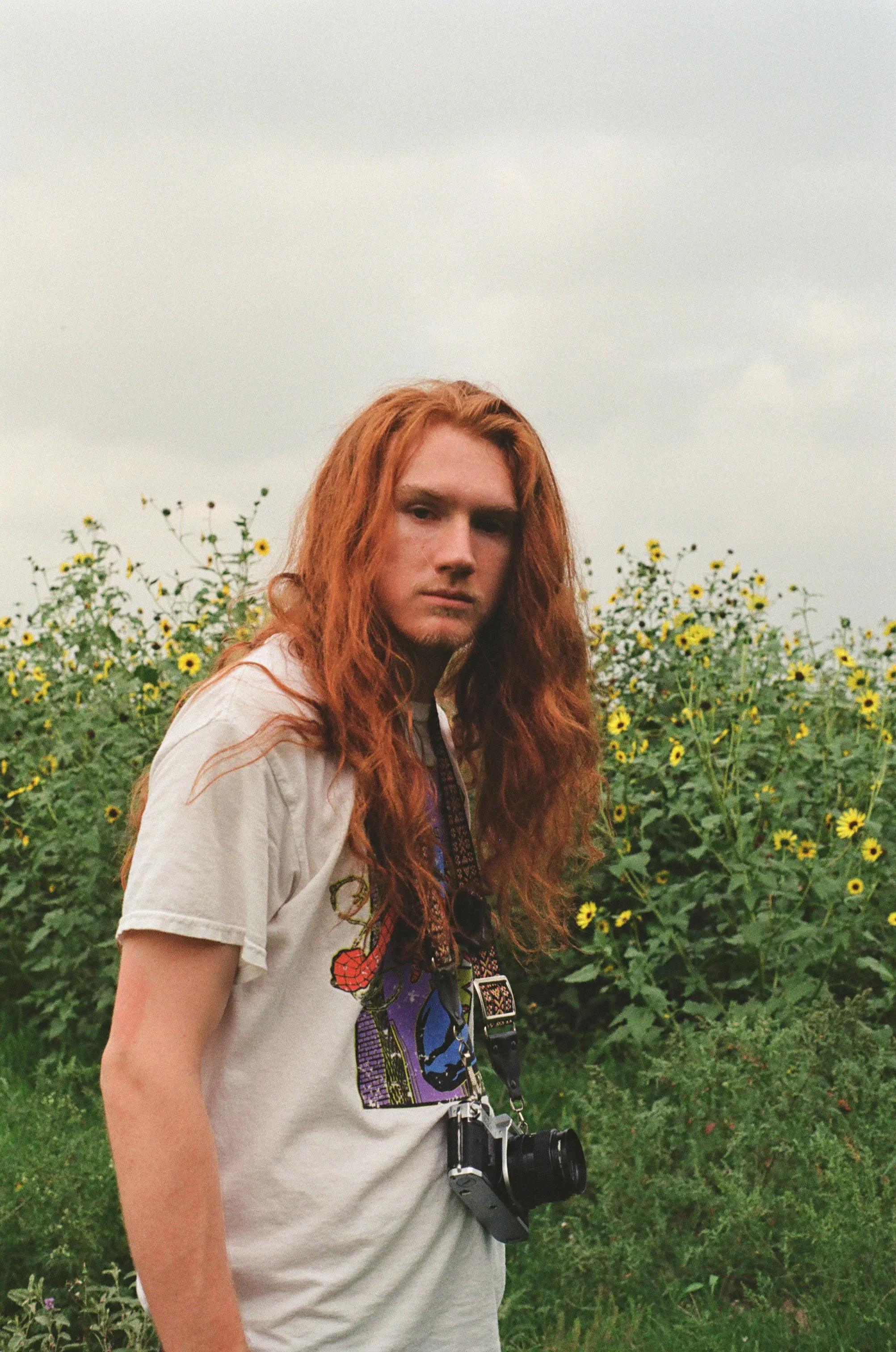 Young woman with long red hair standing in a sunflower field under cloudy sky, wearing a white t-shirt with graphic design and a camera hanging around her neck.