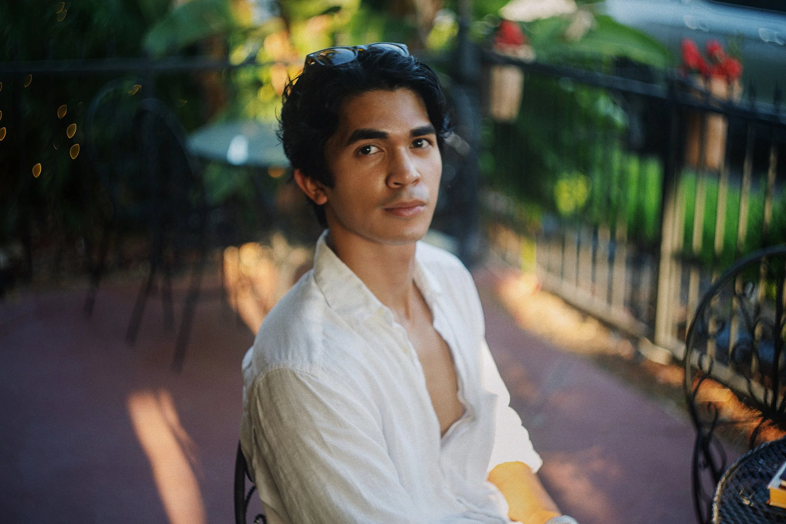 A young man with dark, curly hair and tan skin is sitting outdoors at a patio restaurant. He is wearing a white button-up shirt and looking at the camera with a neutral expression. The background includes a black wrought-iron fence, trees, and patio 