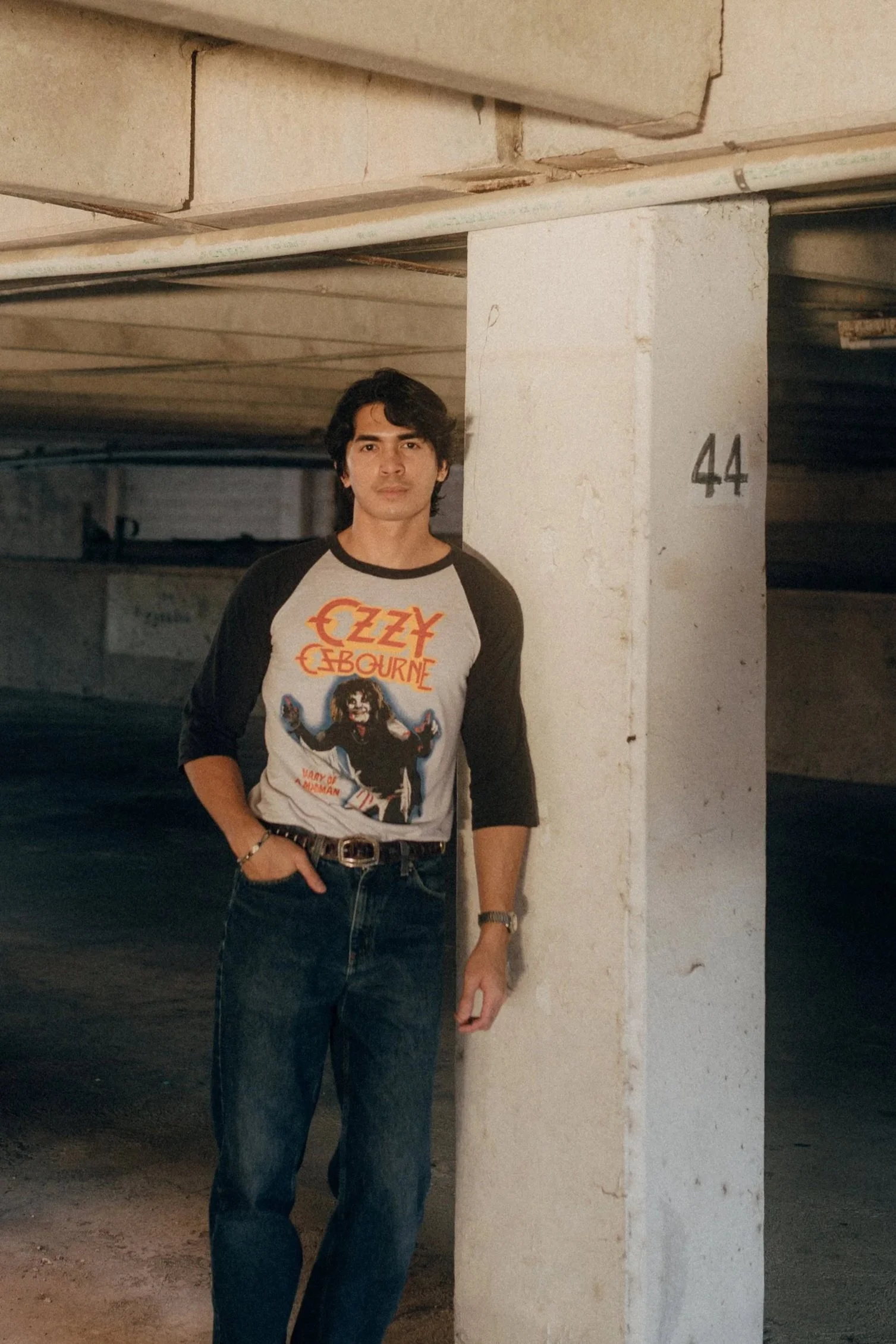 A young man standing next to a concrete pillar in a parking garage, wearing a black and gray Ozzy Osbourne T-shirt and jeans.