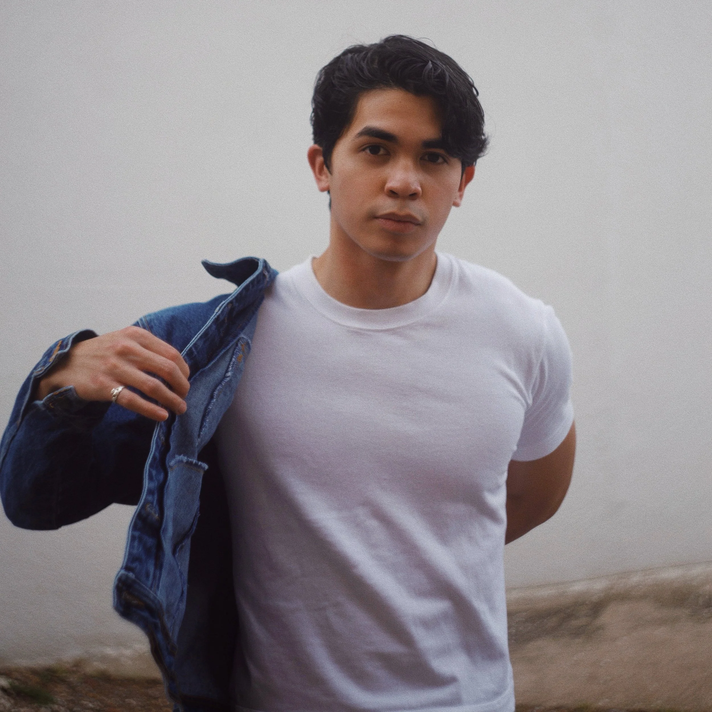 A young man with dark, wavy hair wearing a white t-shirt and a denim jacket over his shoulder, standing outdoors against a plain light sky background.