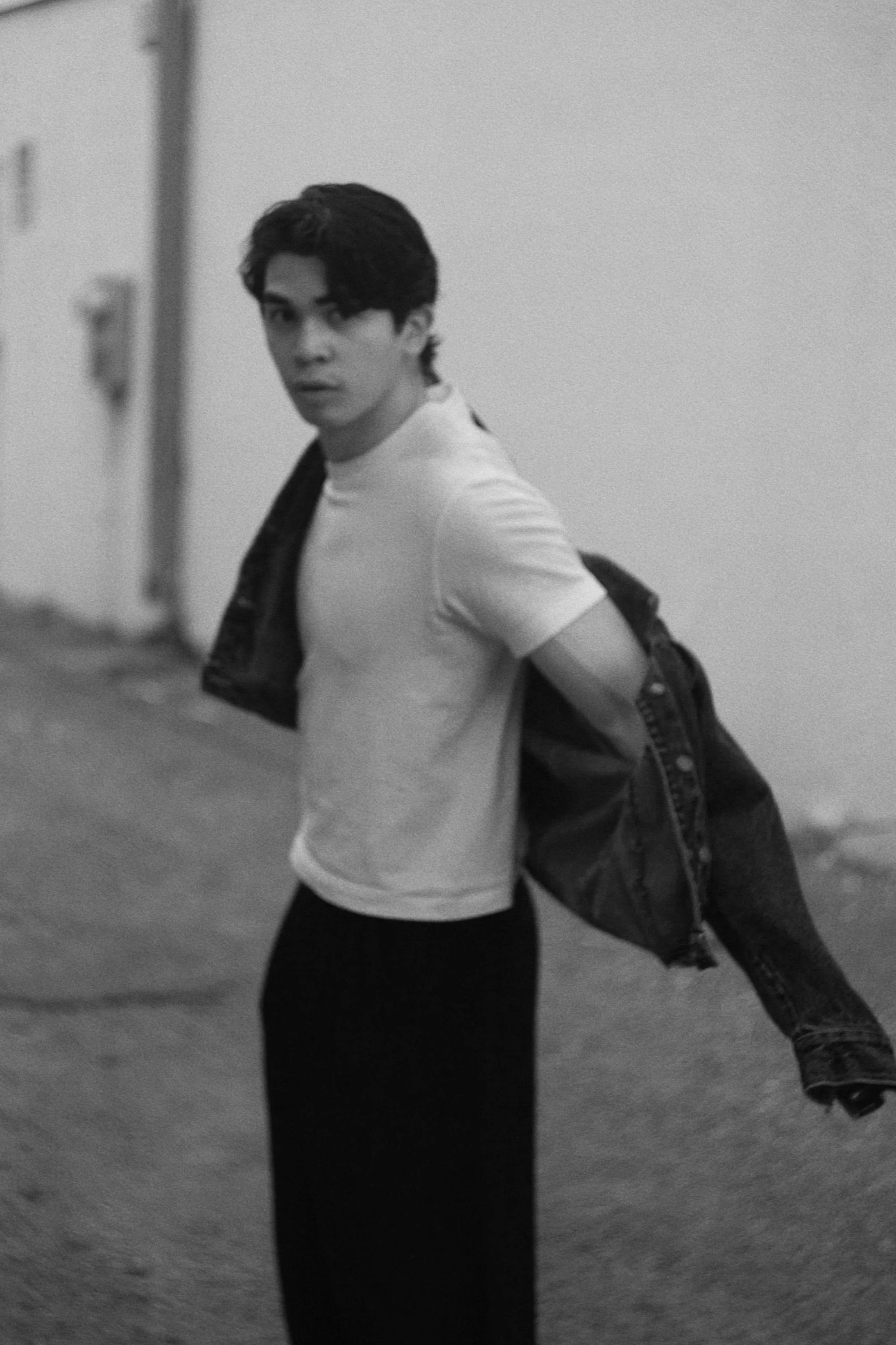 A young man with dark hair, wearing a white t-shirt and black pants, is standing outdoors next to a wall. He is partially removing a denim jacket over his shoulder and looking at the camera.