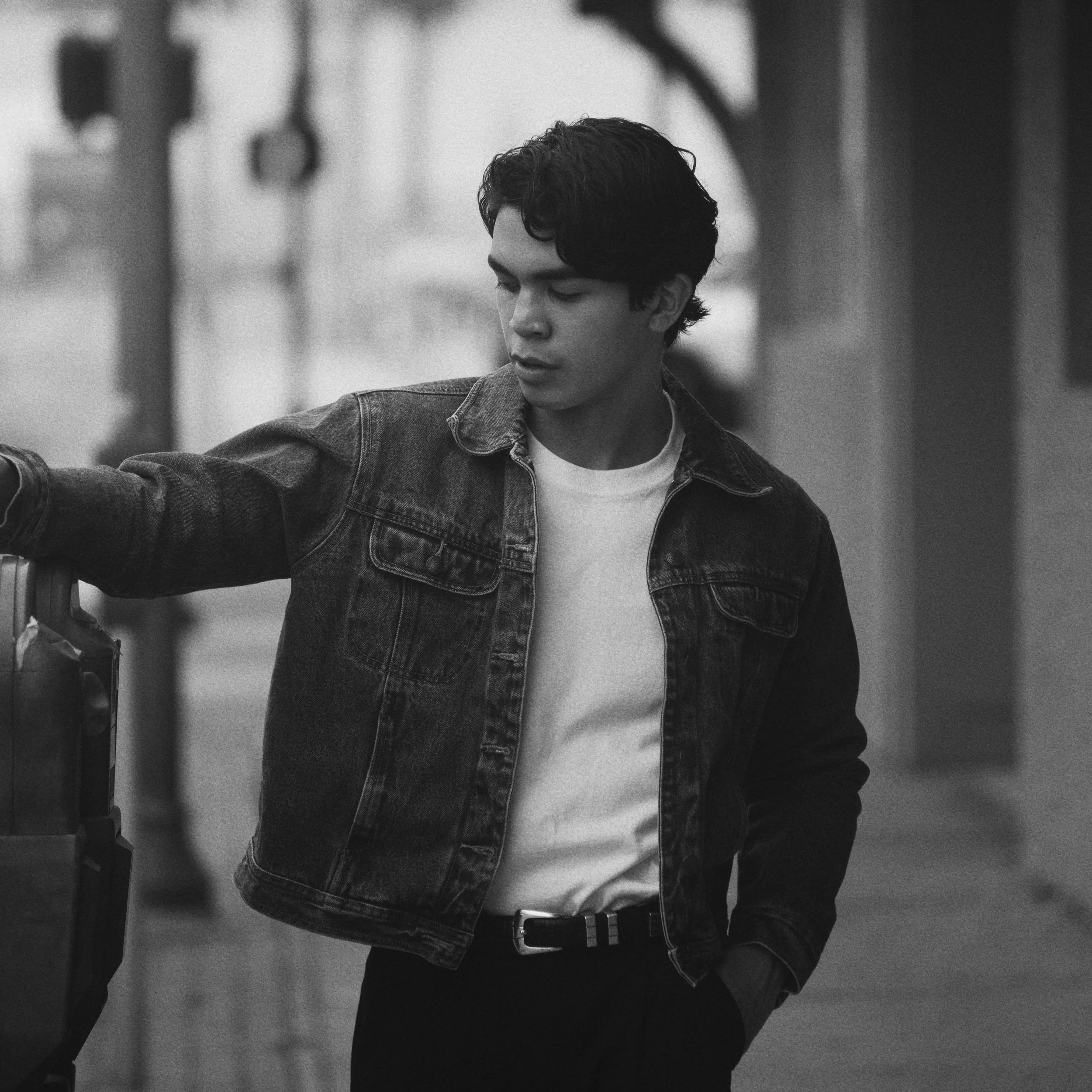 A young man with dark hair, wearing a denim jacket and white t-shirt, standing outdoors and looking down. He has one hand in his pocket and the other resting on a suitcase.