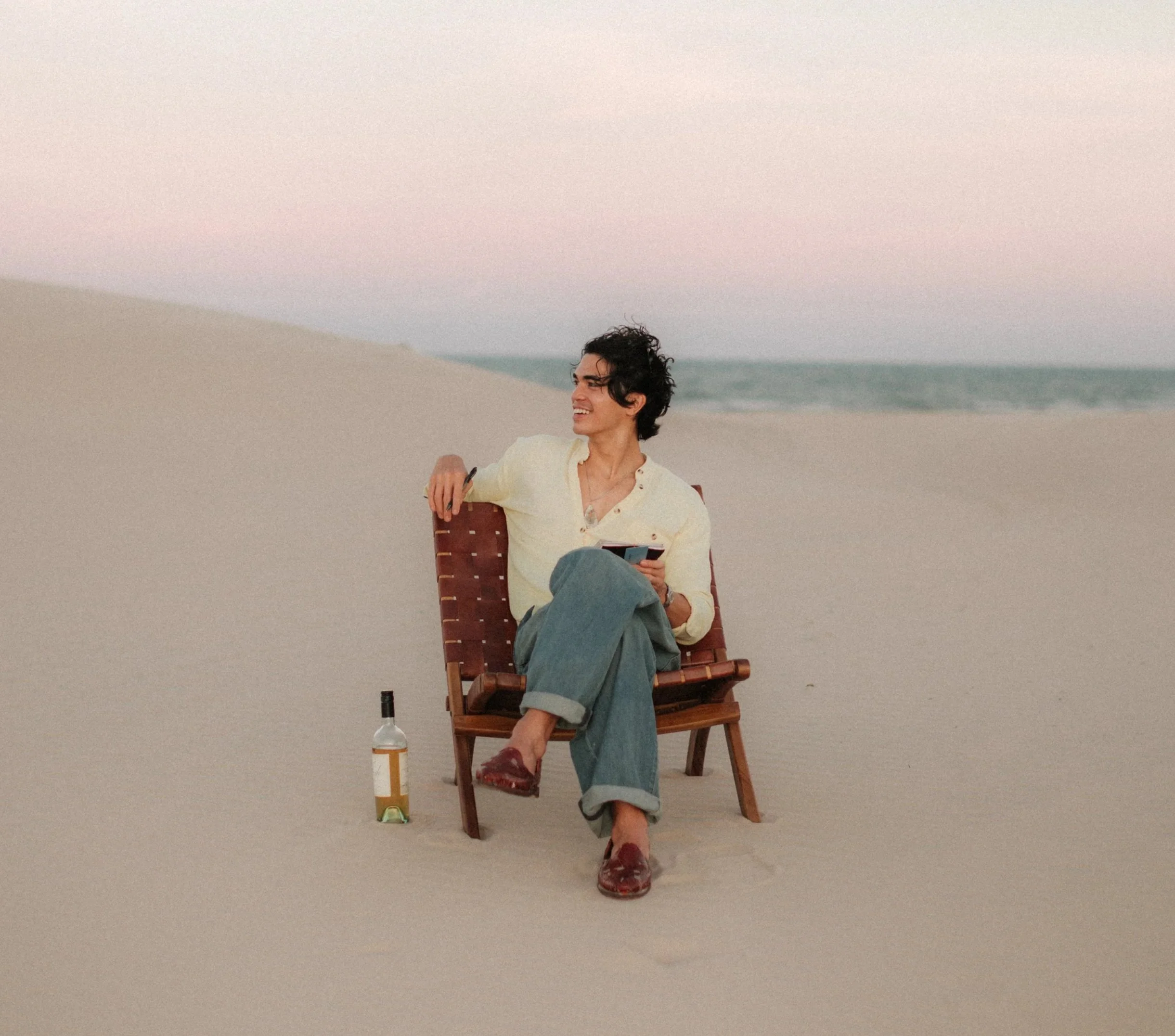 A woman sitting in a wooden chair on a sandy beach with a bottle of wine on the ground nearby. She is smiling, holding a phone, wearing a light yellow shirt, rolled-up jeans, and burgundy shoes, with the ocean and pink sky in the background.