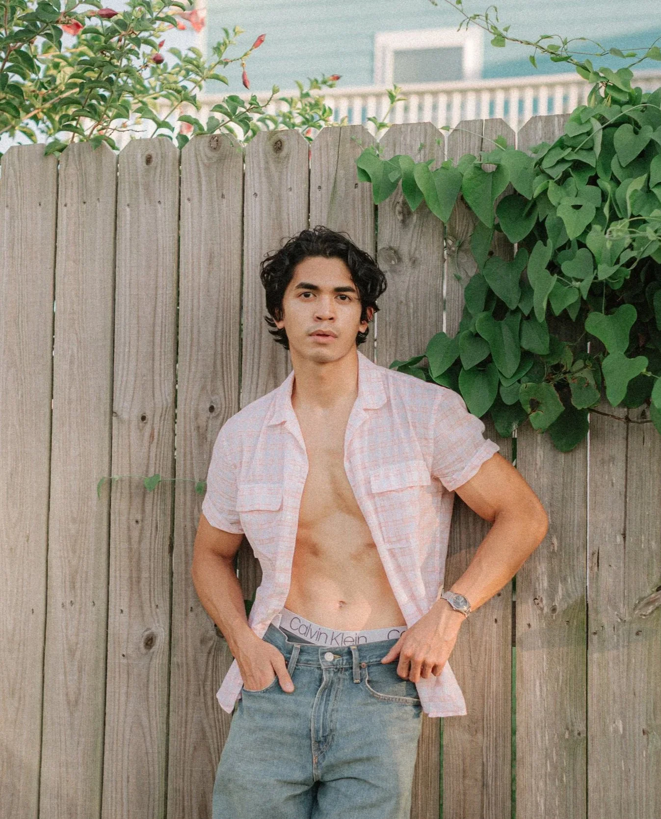 A young man with dark curly hair and light skin standing outdoors in front of a wooden fence, with green leafy plants around him, wearing a partially unbuttoned pink checkered shirt, jeans, and a Calvin Klein waistband visible above his jeans, lookin