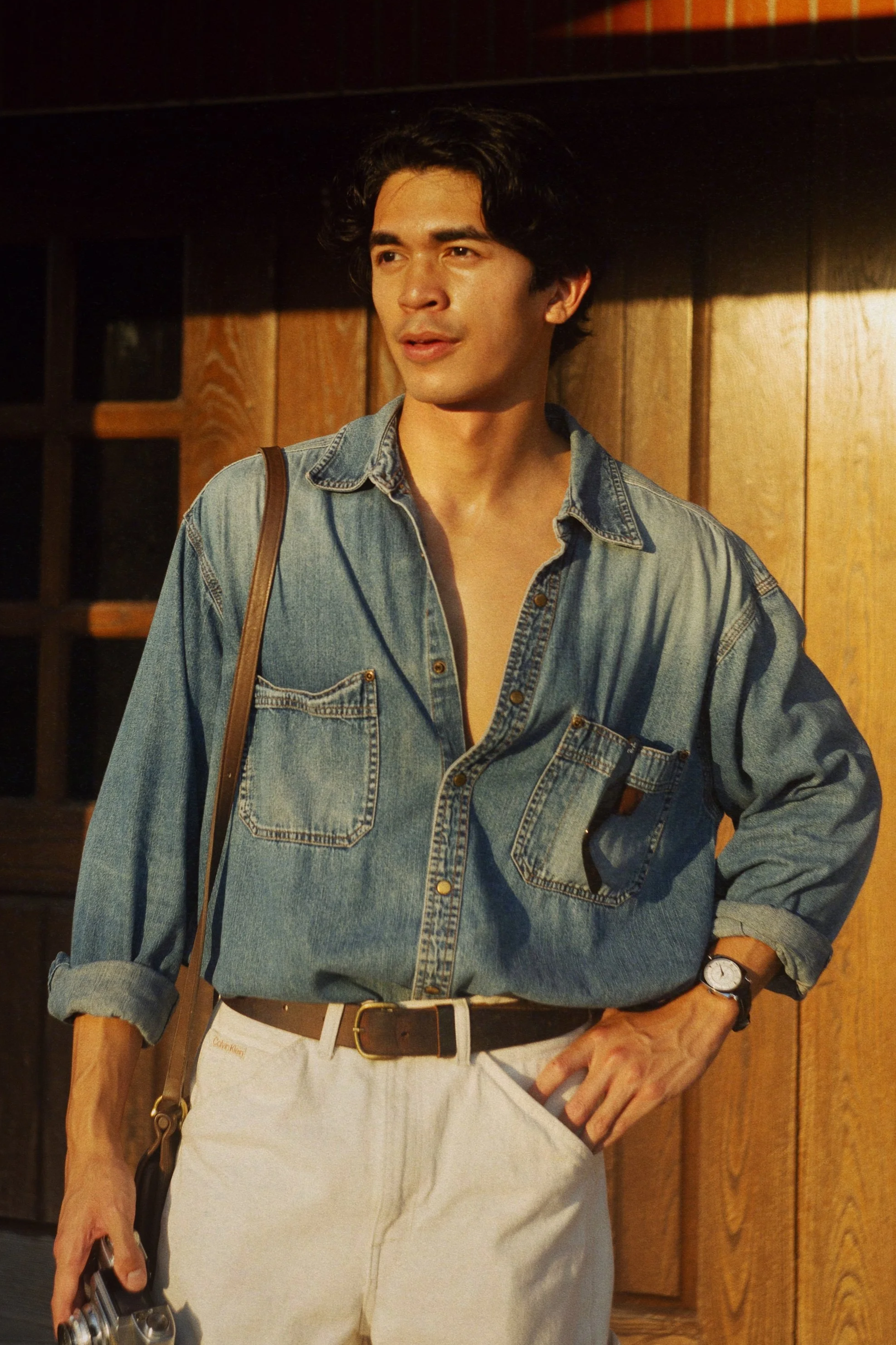 A young man with dark, wavy hair, dressed in a denim shirt with rolled-up sleeves, white pants, a black belt, and a watch, stands beside a wooden wall, holding a camera in his right hand.