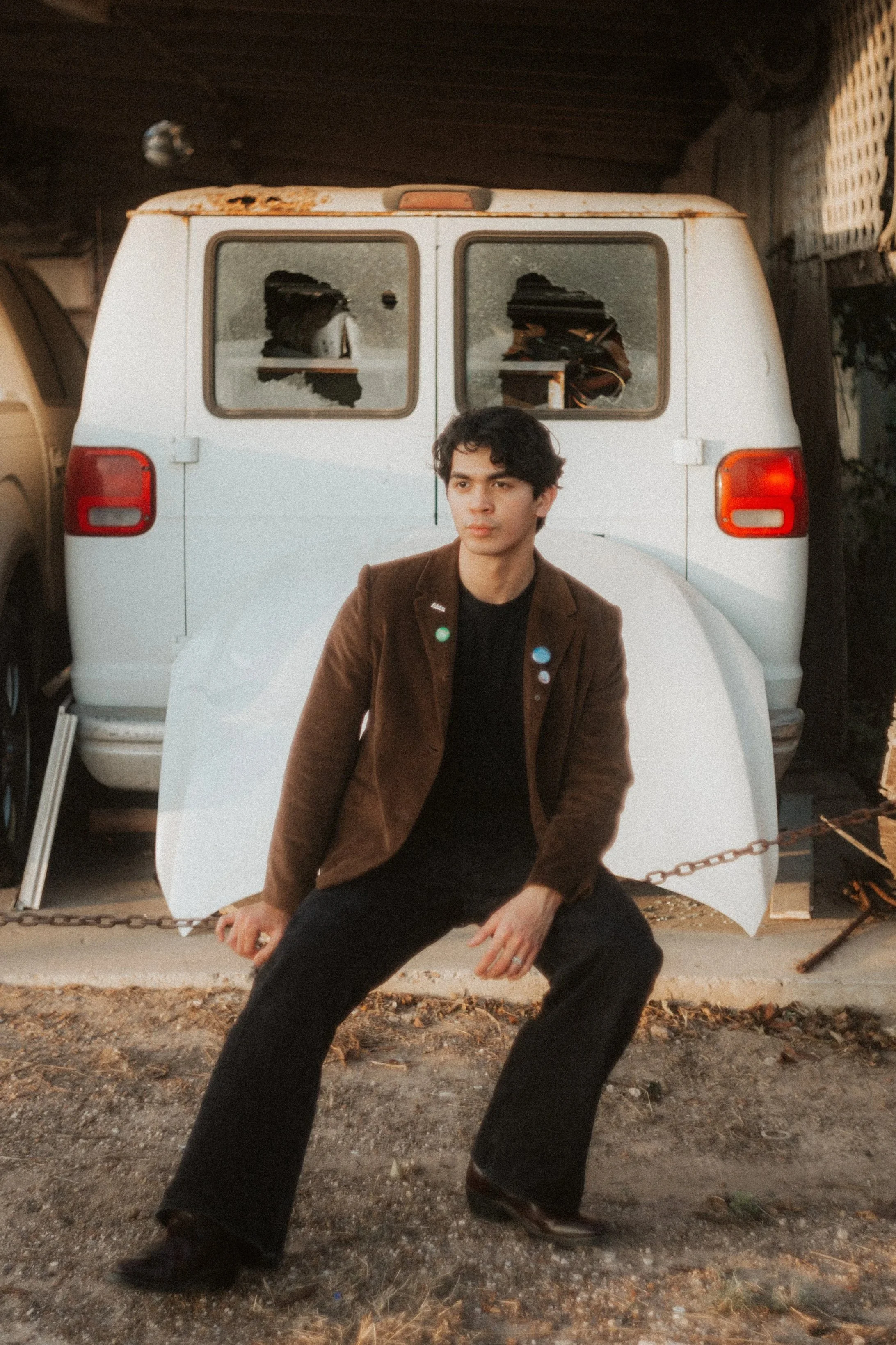 A young man sitting in front of a white van, wearing a brown jacket and black pants, outdoors during sunset.