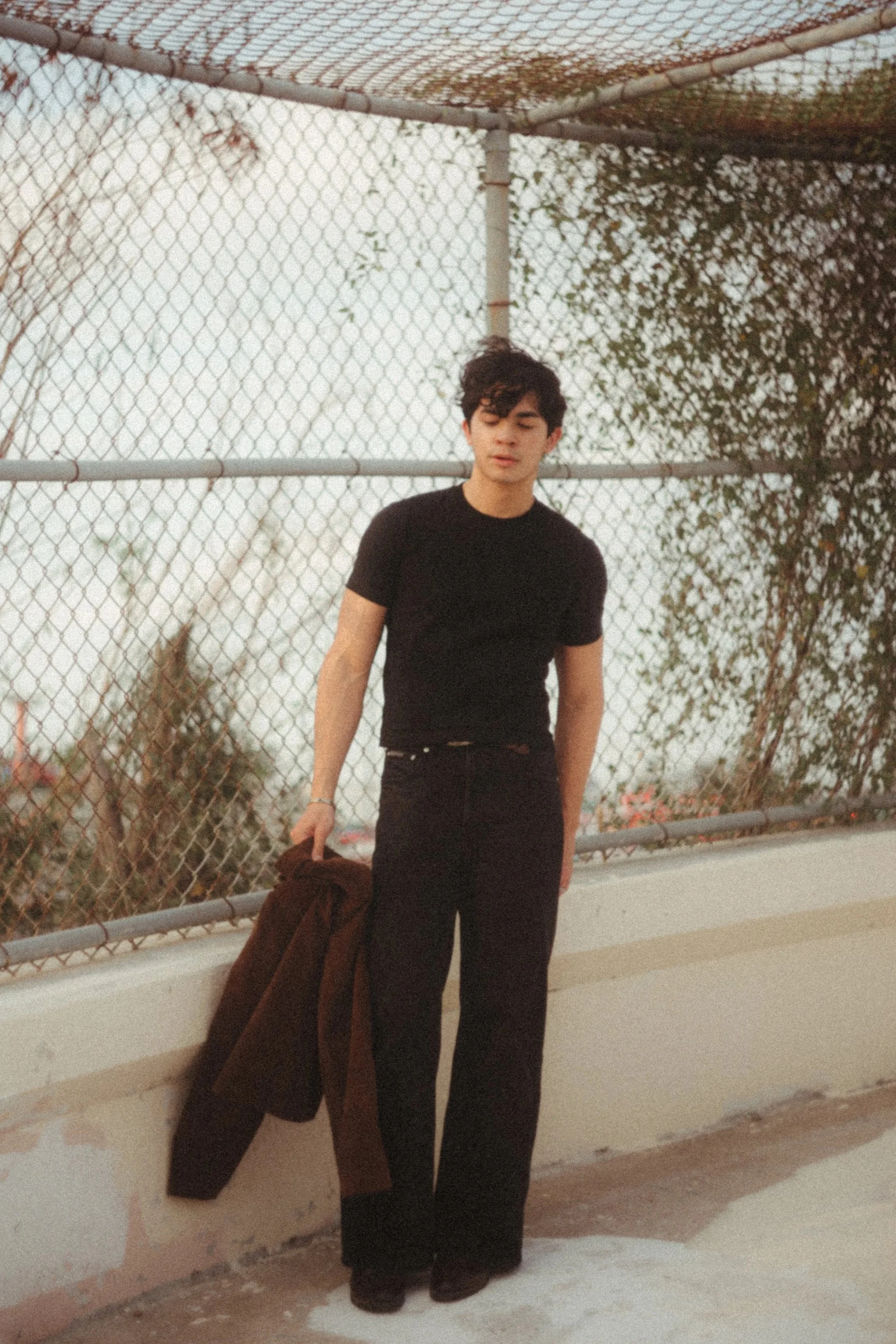 A young man with dark curly hair wearing a black t-shirt and black pants stands next to a white concrete wall with a brown coat draped over it, against a background of a chain-link fence and trees.
