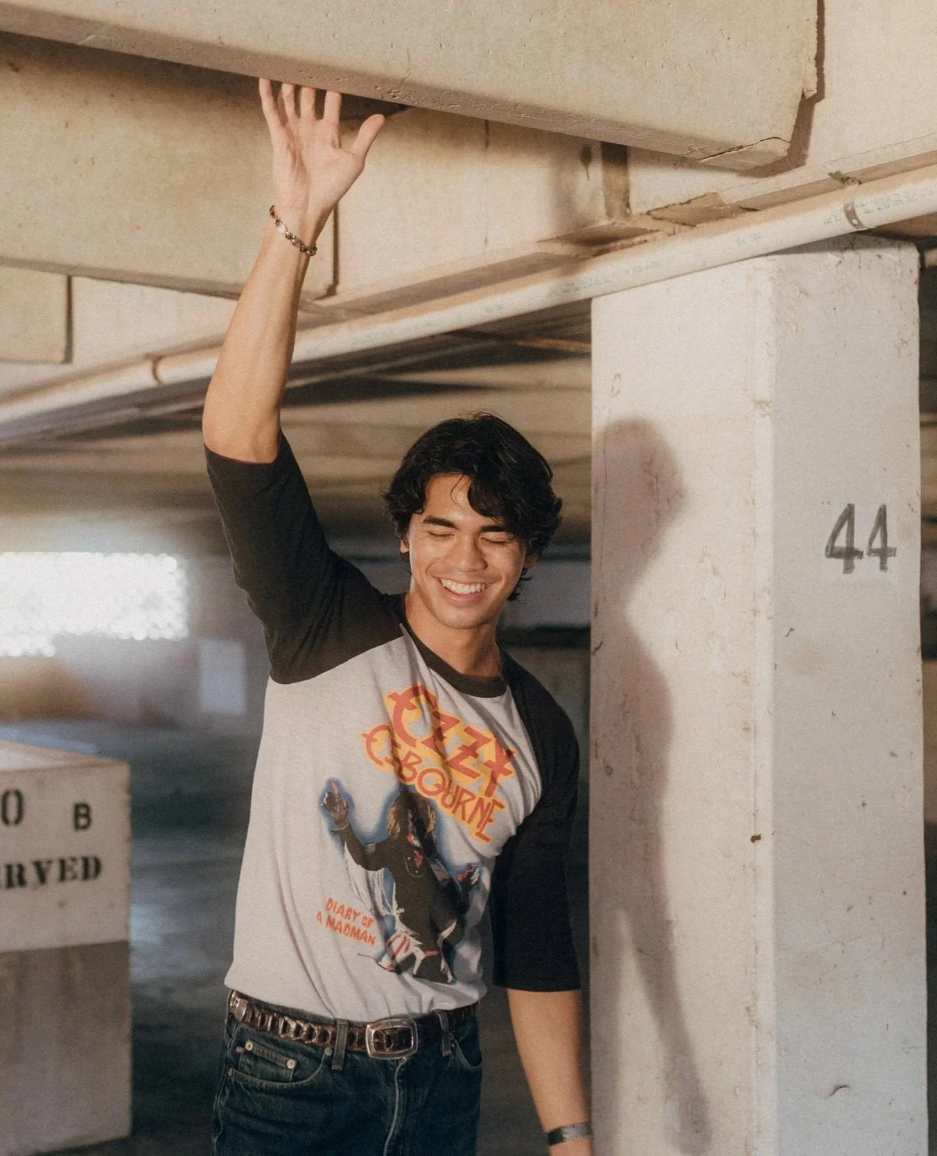 A young man with dark hair smiling and wearing a 'Ozzy Osbourne' T-shirt, standing in a parking garage, with his hand raised touching the ceiling.