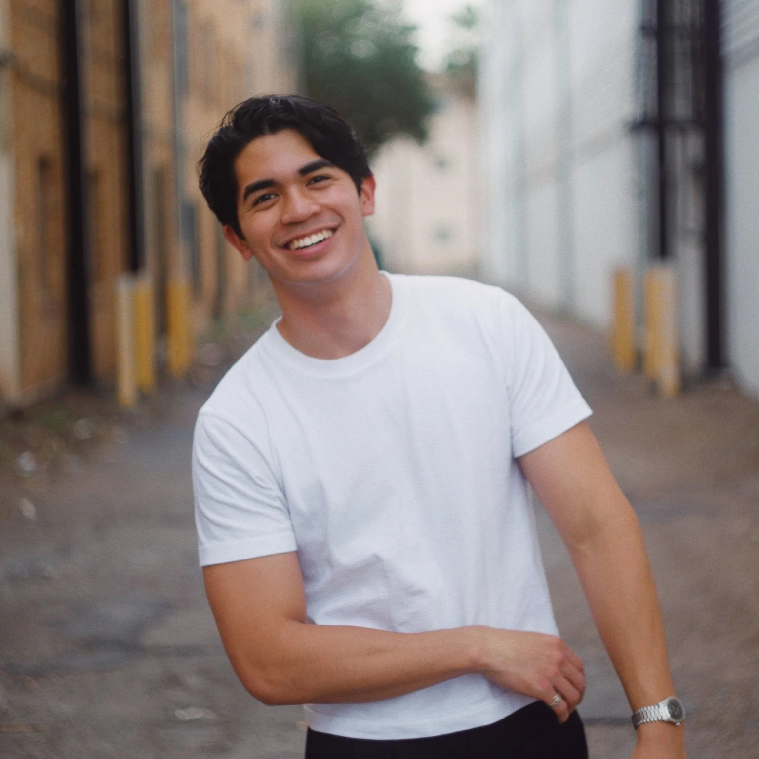 A young man with dark hair smiling outdoors, wearing a white t-shirt and a watch, standing on a street with industrial and wooden buildings on either side.