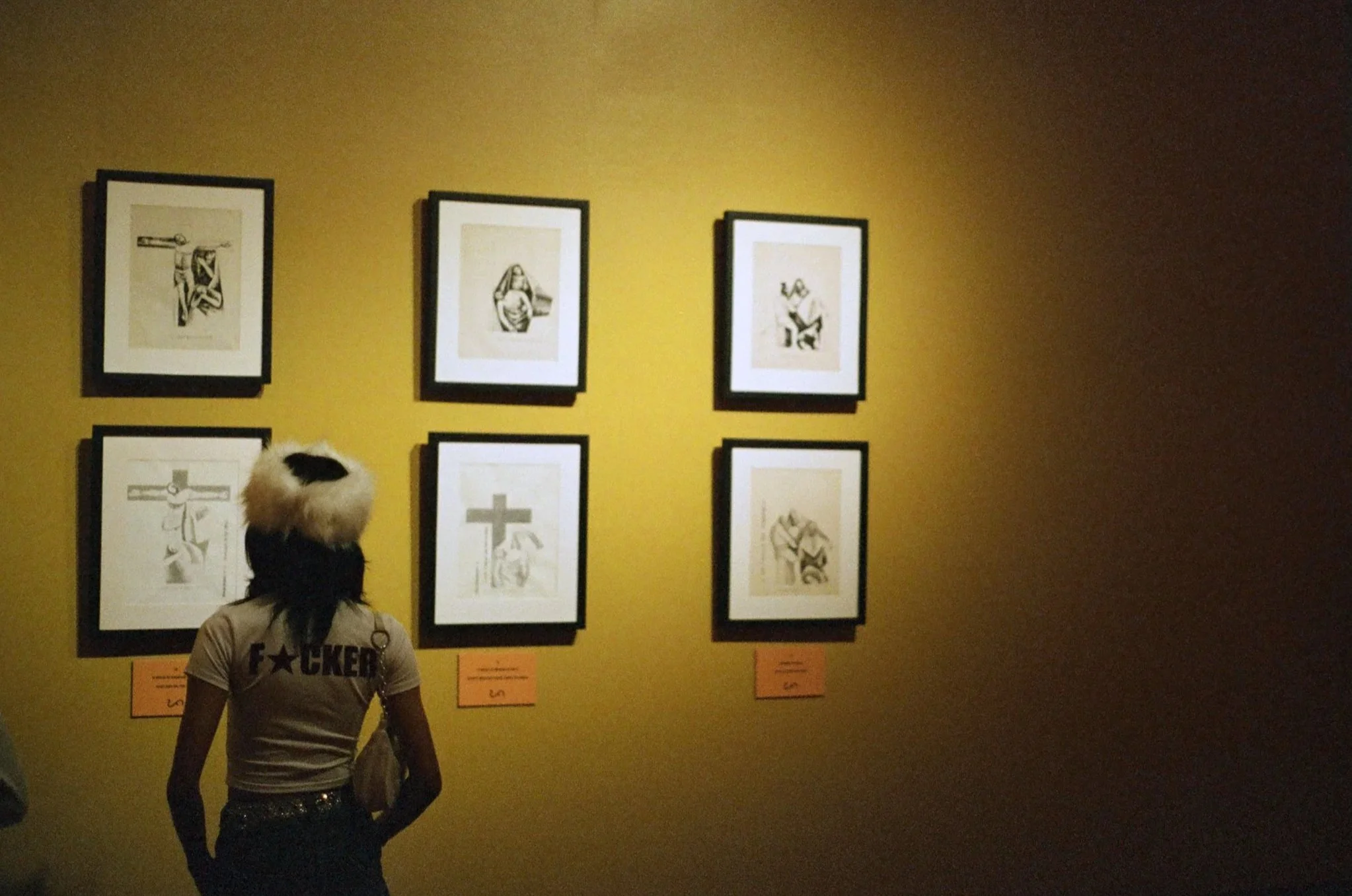 A person with a fur hat and a shirt that says 'F**KER' looks at six framed black-and-white illustrations of the crucifixion on a yellow gallery wall, with numbered orange labels below.