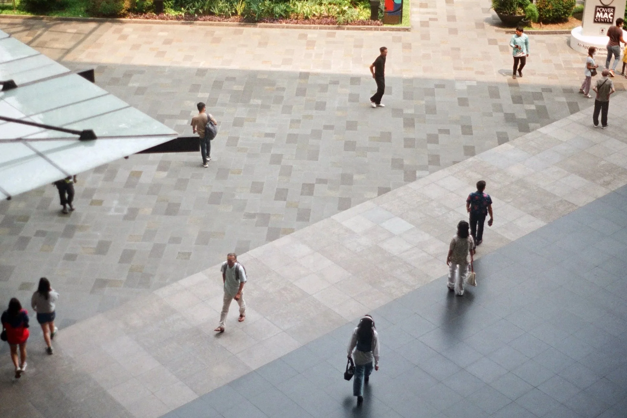 People walking in an outdoor shopping or public area, with trees and planters, and a large sign reading 'Power Mac Center' in the background.