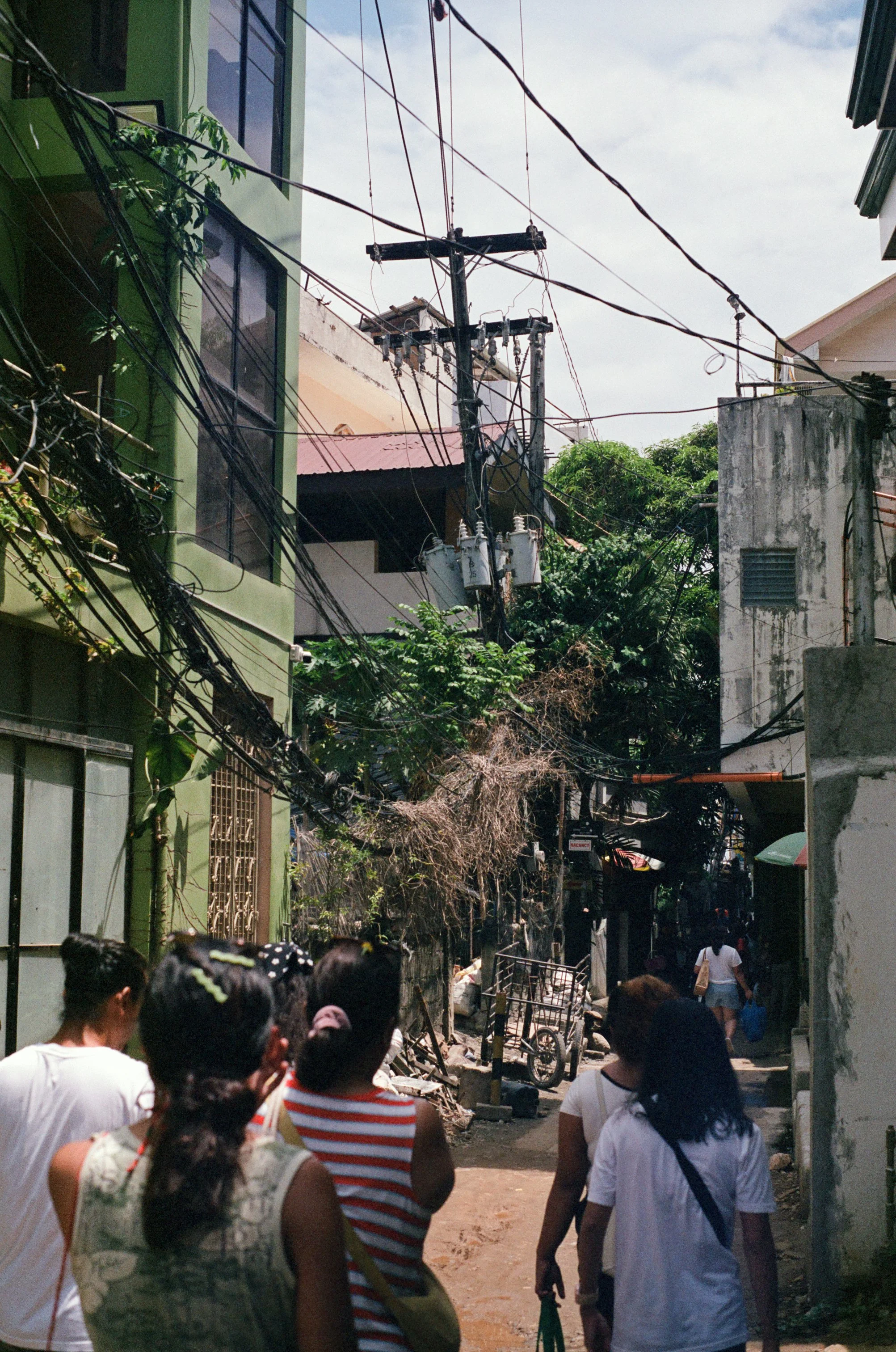 A crowded alleyway in an urban area with multiple pedestrians walking. Overhead, tangled power lines and utility poles crisscross above narrow streets lined with buildings, some with visible damage or wear.