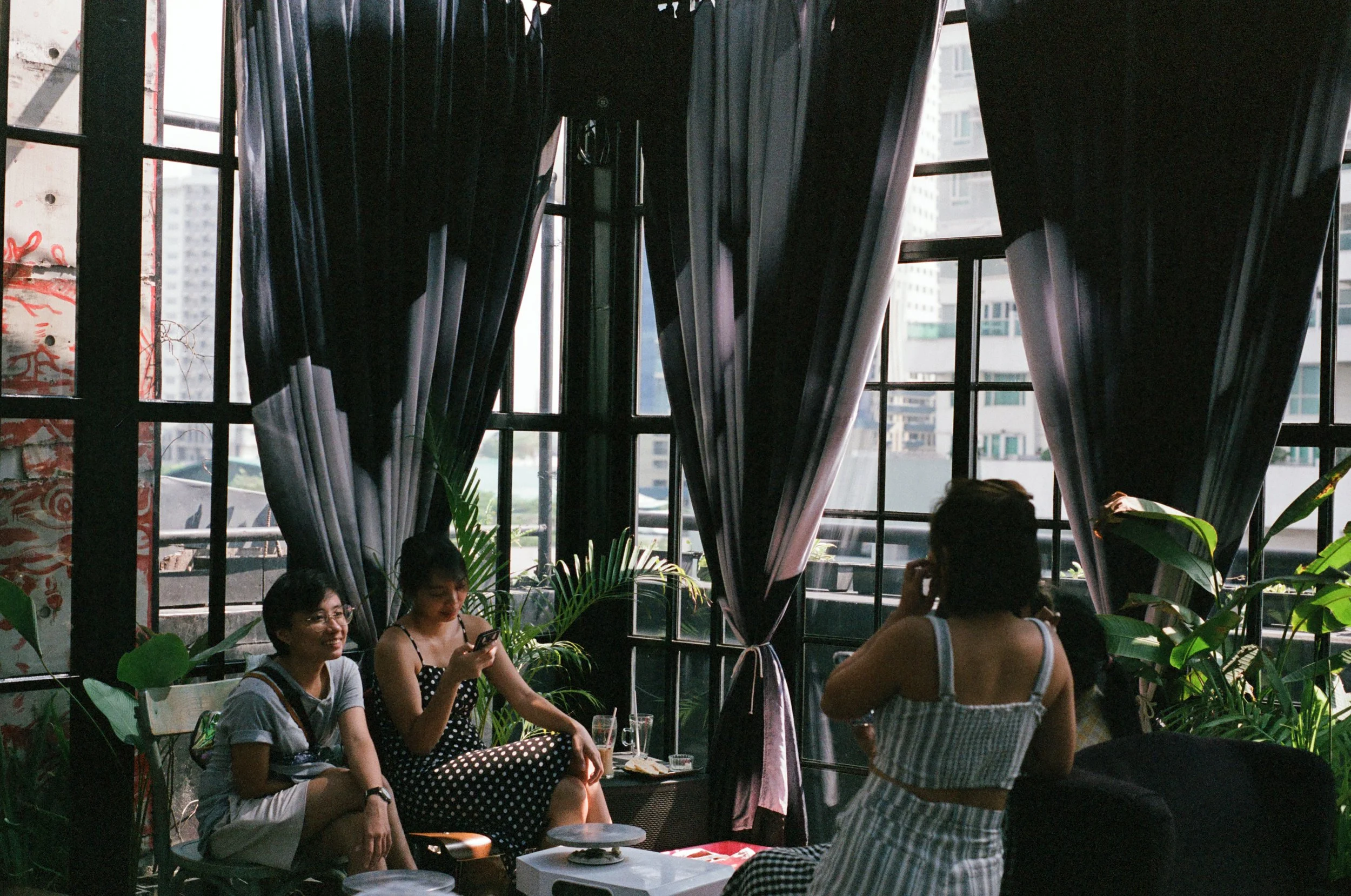 Three people sitting and chatting in a cozy indoor space with large windows and black curtains, with city buildings visible outside, and green plants around.