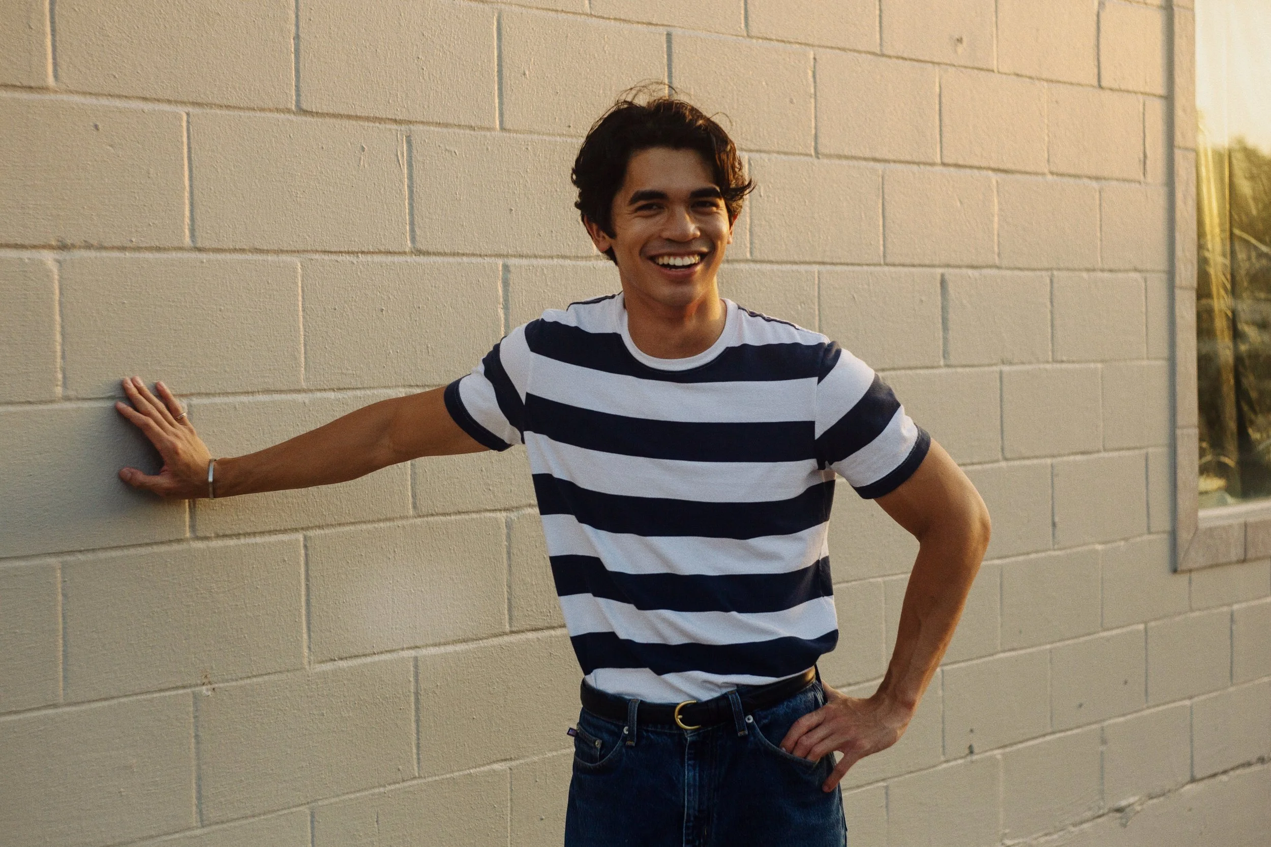 A young man with dark hair in a striped T-shirt smiling and standing outdoors near a beige brick wall, with one hand on the wall and the other on his hip.