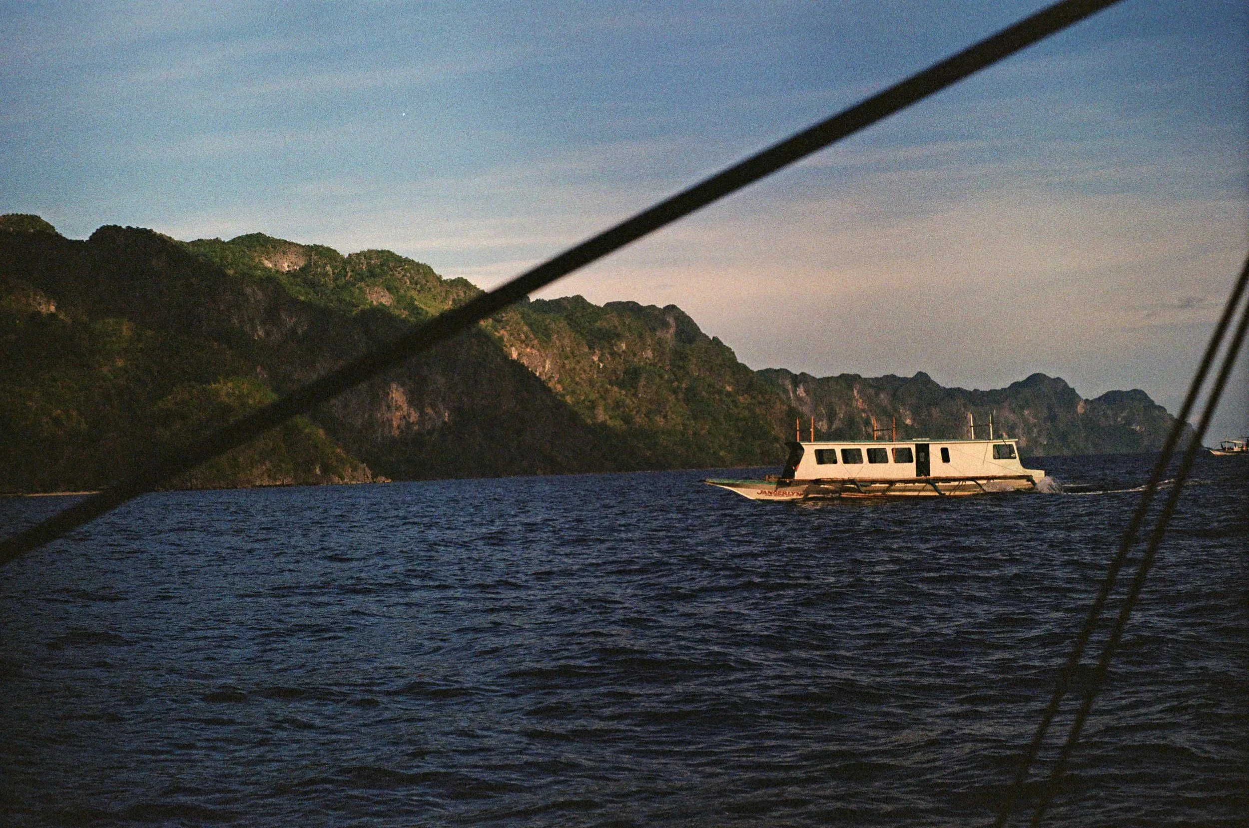 A boat sailing on dark blue water near a green mountainous coastline during dusk or dawn.