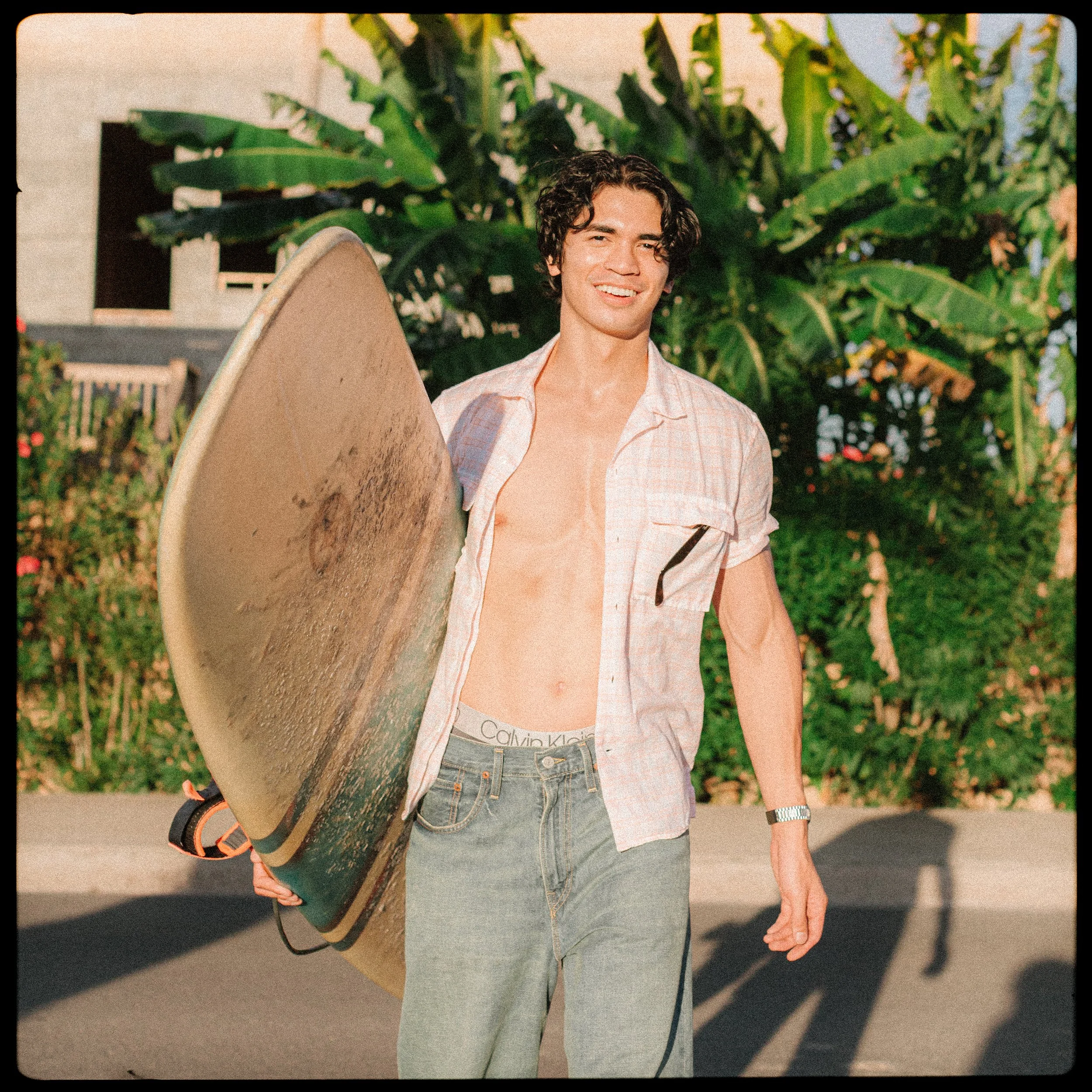 A young man with curly dark hair, shirt open showing bare chest, holding a surfboard, standing outdoors with lush green plants in the background.