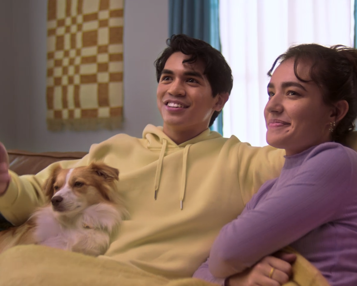 A young man and woman sitting on a couch, smiling and relaxing with a dog on the man's chest in a cozy living room.