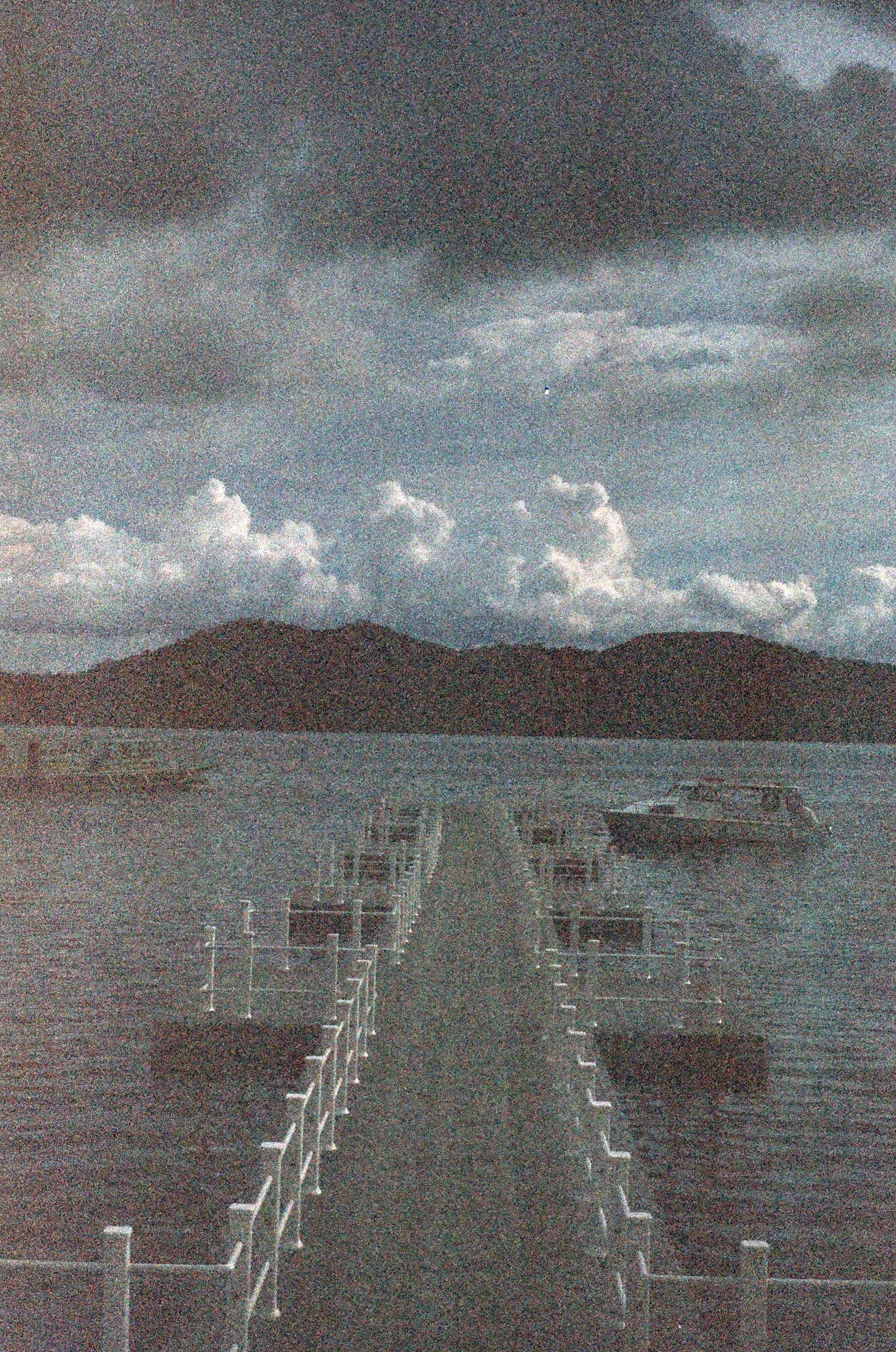 A dock extending into a lake with boats anchored along the sides, mountains in the background, and a cloudy sky.
