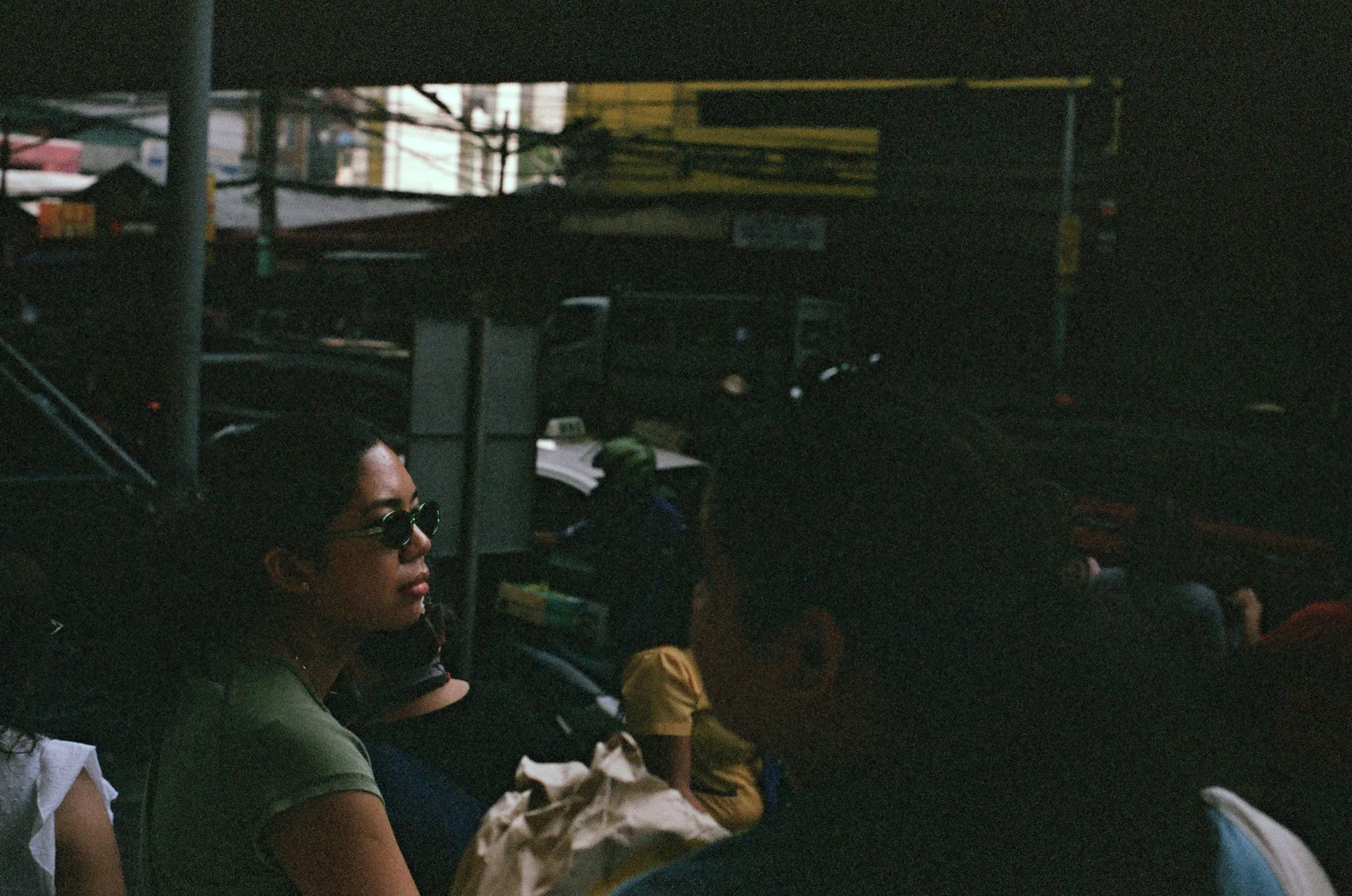 A woman wearing sunglasses and a green shirt sitting outdoors during daytime, other people around her, and a busy urban background.