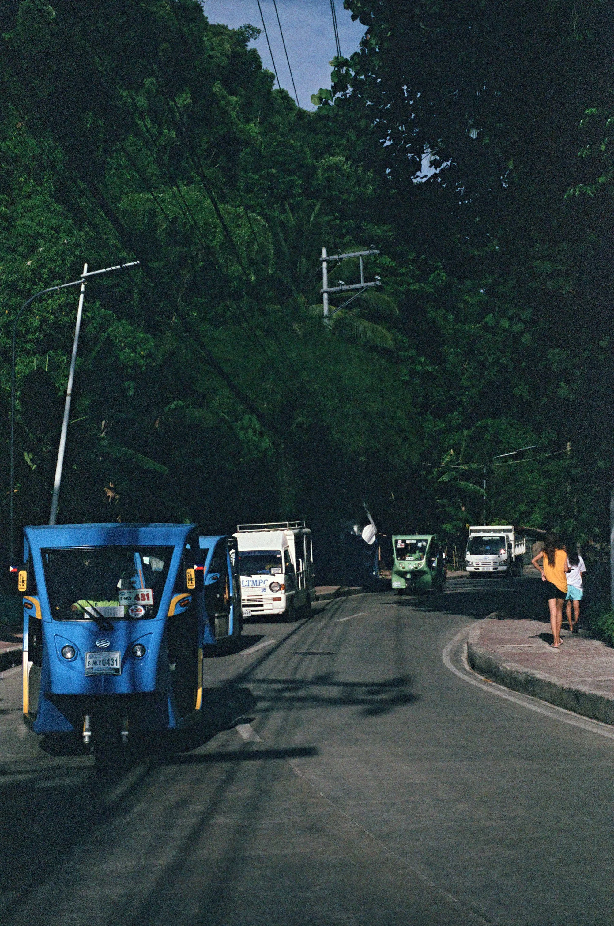 Nighttime street scene with parked vehicles, including a blue electric rickshaw, surrounded by lush green trees, and two women walking on the sidewalk.